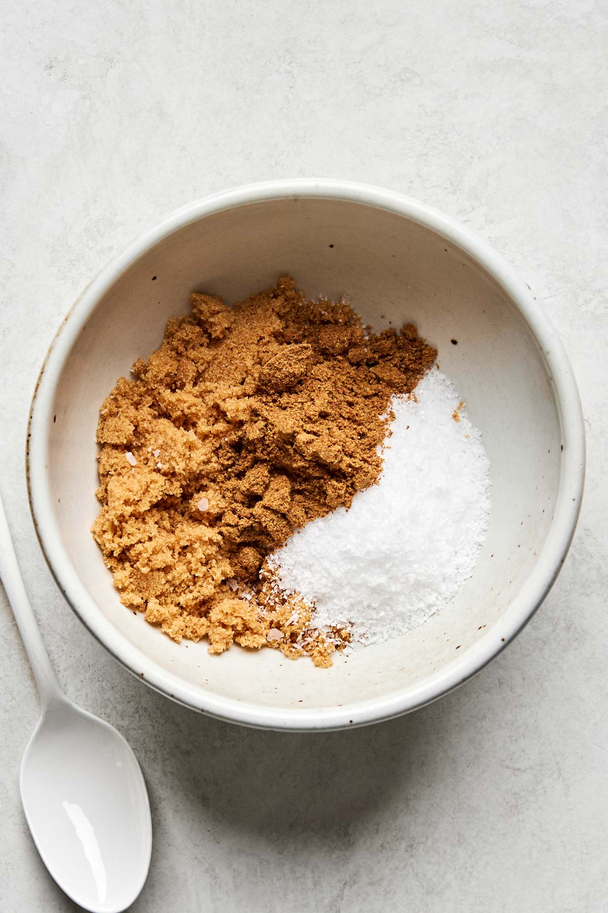 A white bowl containing brown sugar and coarse salt, placed on a light surface next to a white spoon.