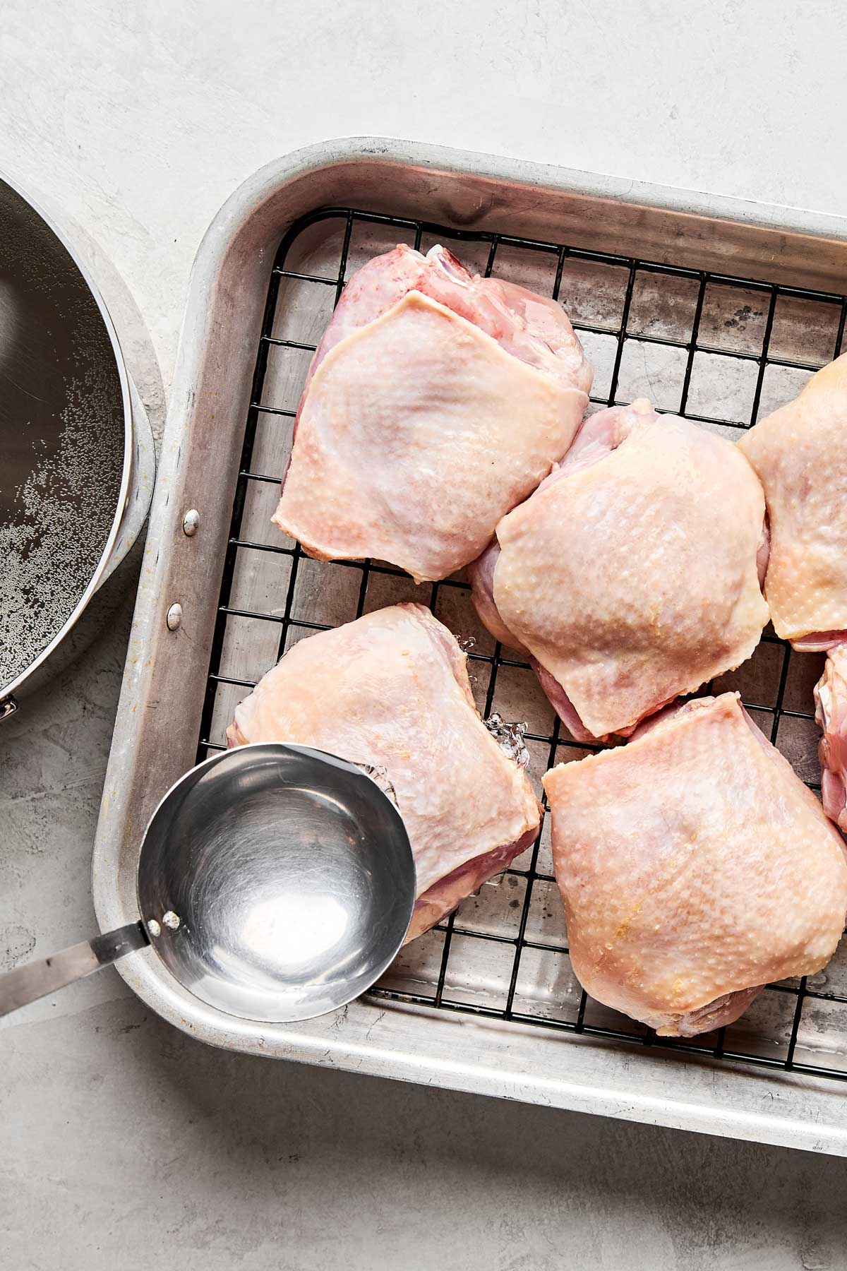 Six raw chicken thighs on a wire rack set inside a baking tray, with a ladle of water next to them and a pan partially visible on the side, all placed on a light-colored surface.