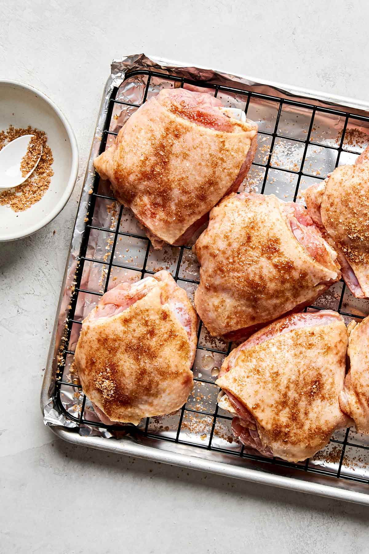 Raw, seasoned chicken thighs arranged on a wire rack over a foil-lined baking sheet, with a bowl of brown seasoning mixture and a spoon nearby on a light countertop.