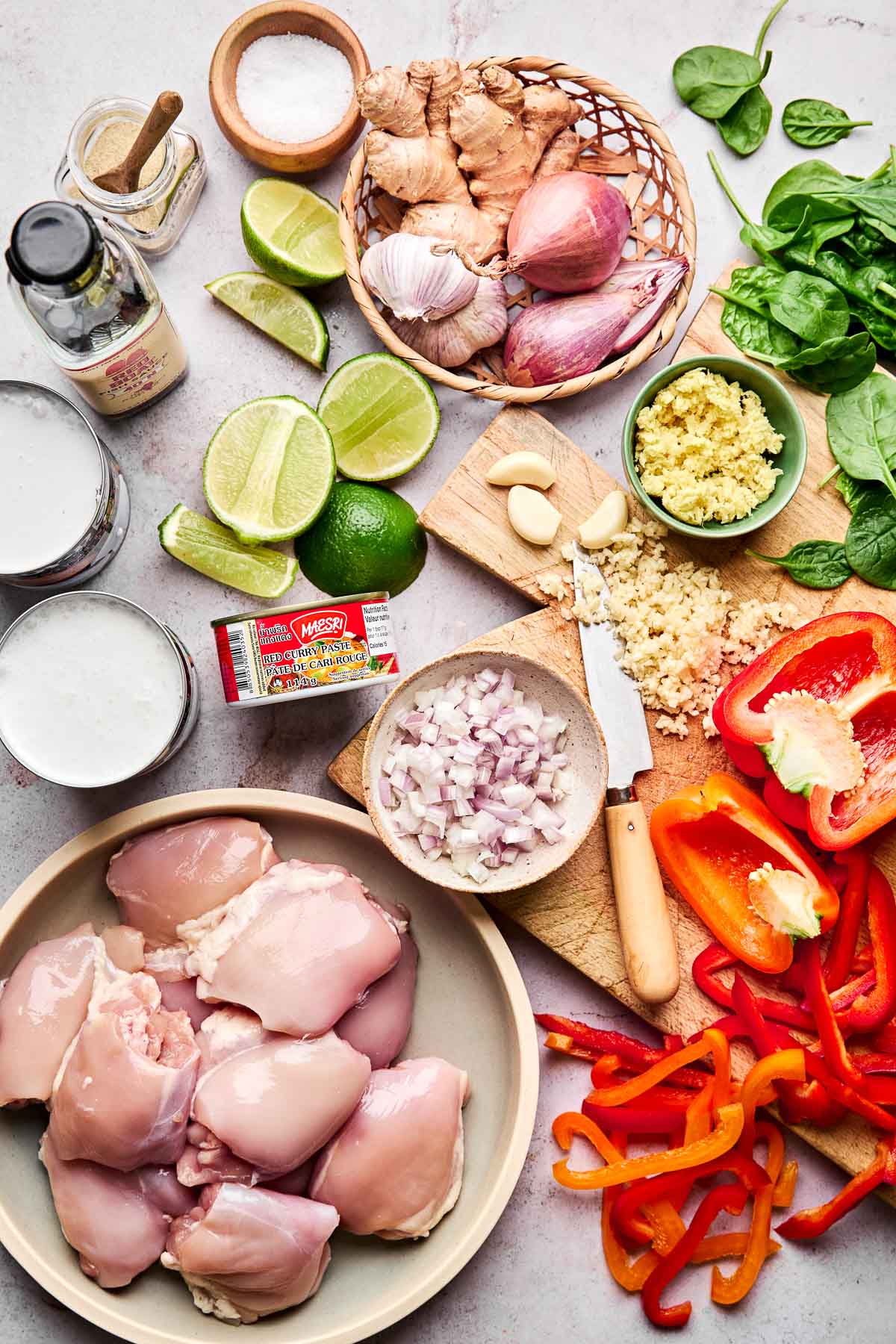 An overhead view of raw chicken thighs, sliced red bell peppers, chopped shallots, garlic, ginger, lime halves, spinach, coconut milk, and seasonings arranged on a light countertop.