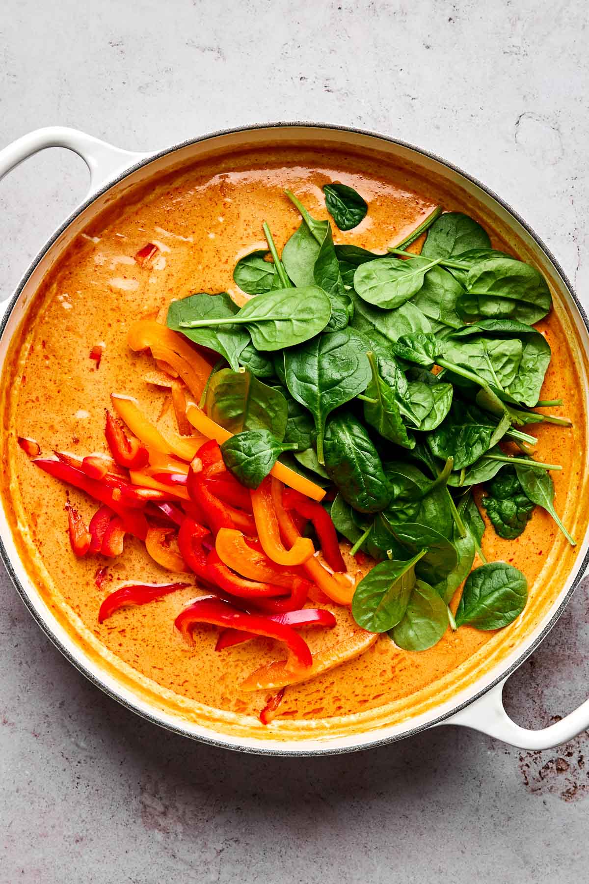 A white pan filled with creamy orange curry sauce, topped with fresh spinach leaves and sliced red and yellow bell peppers, on a light gray surface.