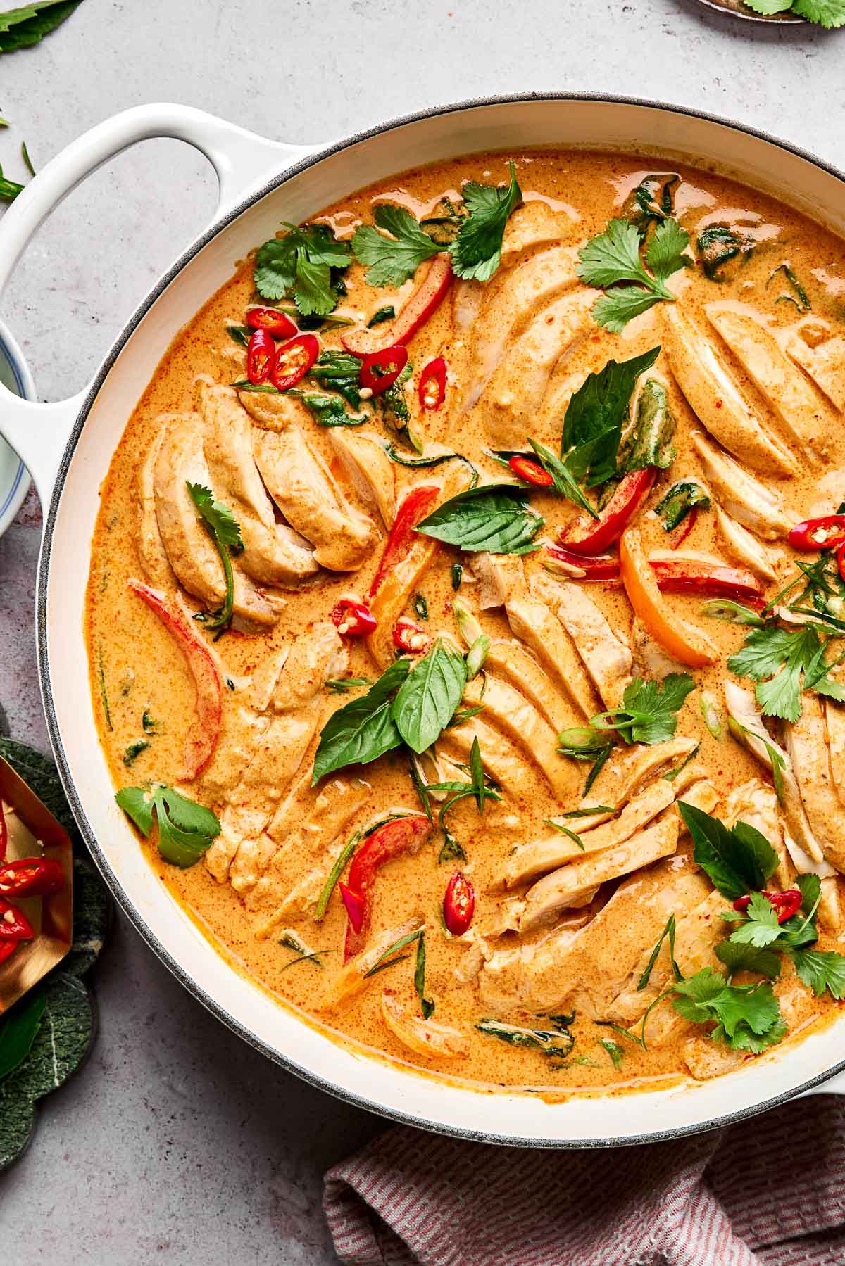 A white pan filled with sliced chicken in a creamy orange curry sauce, garnished with fresh herbs, red chili slices, and red bell peppers. The dish is on a light-colored surface with a striped cloth nearby.