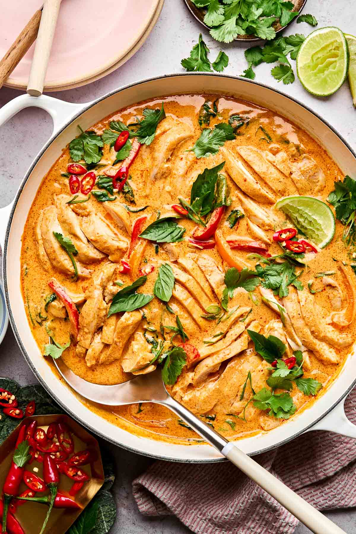 A white pot filled with creamy orange Thai curry, sliced chicken, red chili, fresh herbs, and lime wedges. Garnishes and ingredients like cilantro, lime, and chili are scattered around the pot on a light surface.