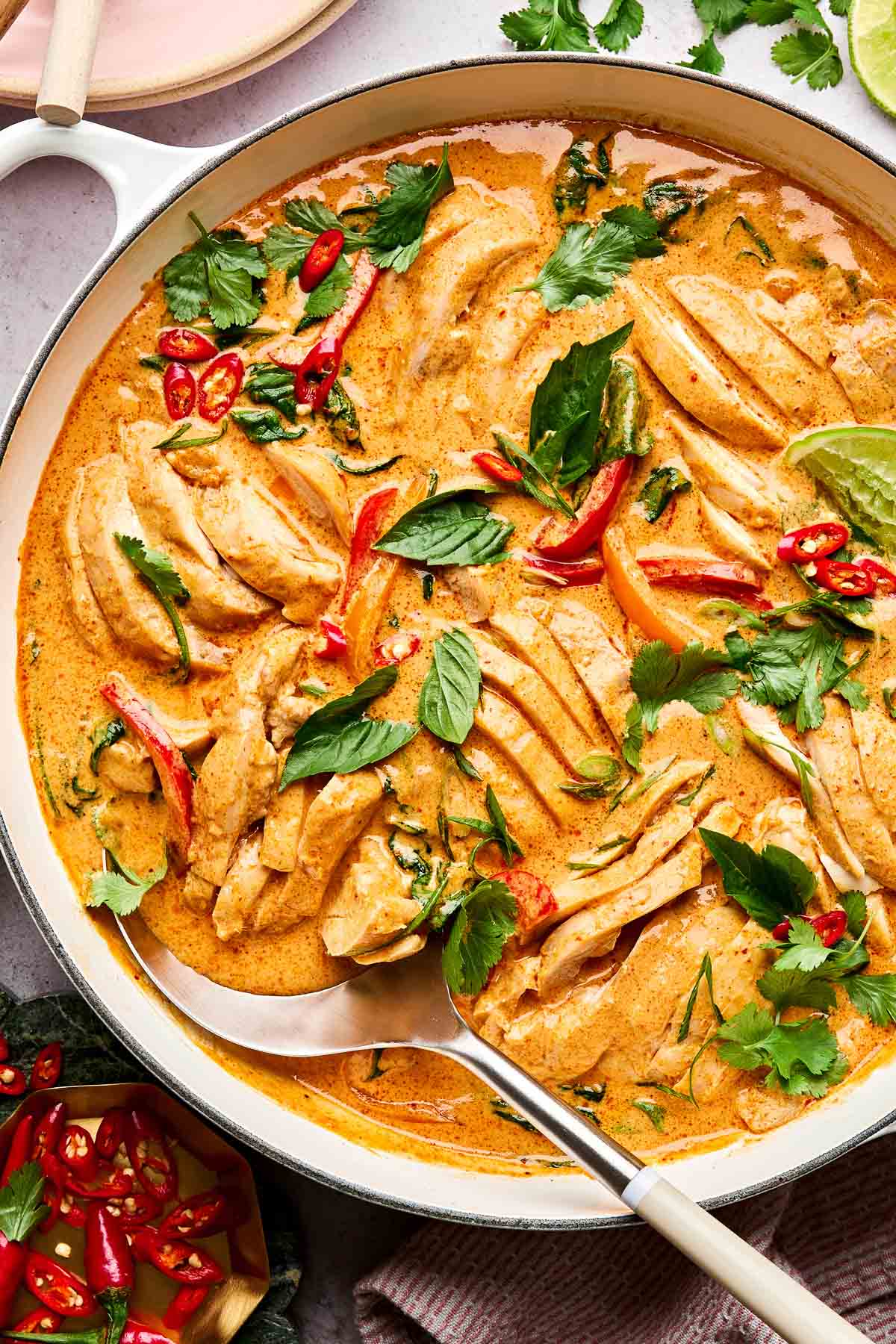 A pot of creamy Thai red curry with sliced chicken, garnished with fresh herbs, red chili slices, and lime wedges. A serving spoon rests in the curry, and fresh cilantro and chili are scattered nearby.