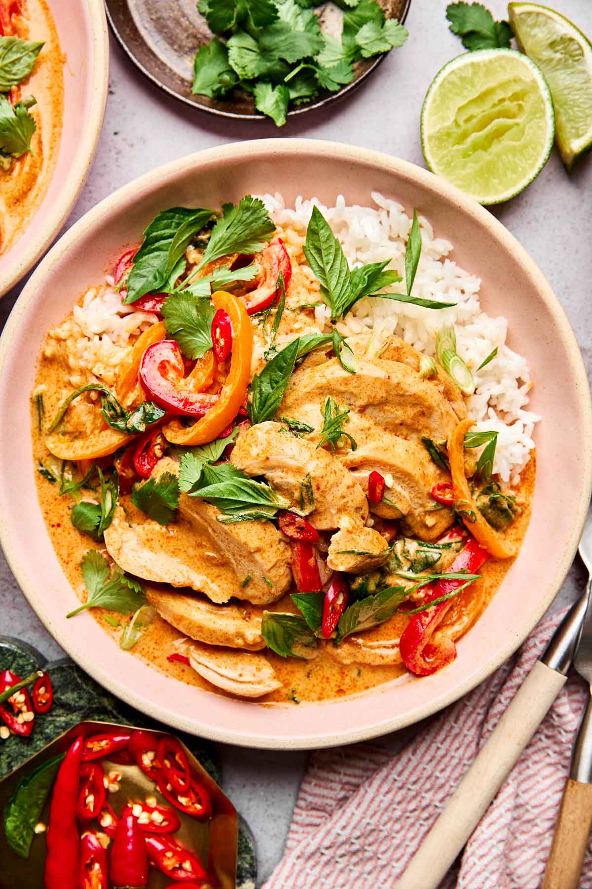 A plate of Thai red curry with chicken, bell peppers, herbs, and red chili slices served over white rice, garnished with fresh cilantro and basil. Lime wedges and more herbs are visible beside the dish.