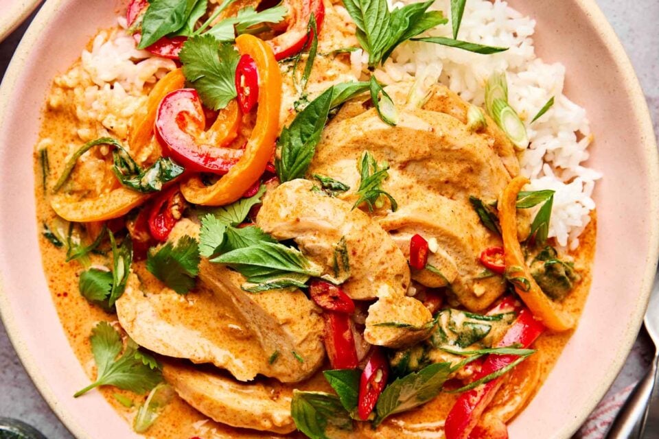 A plate of Thai red curry with sliced chicken, red and yellow bell peppers, fresh herbs, and white rice. The dish has a creamy, orange-colored sauce and is garnished with cilantro and basil leaves.