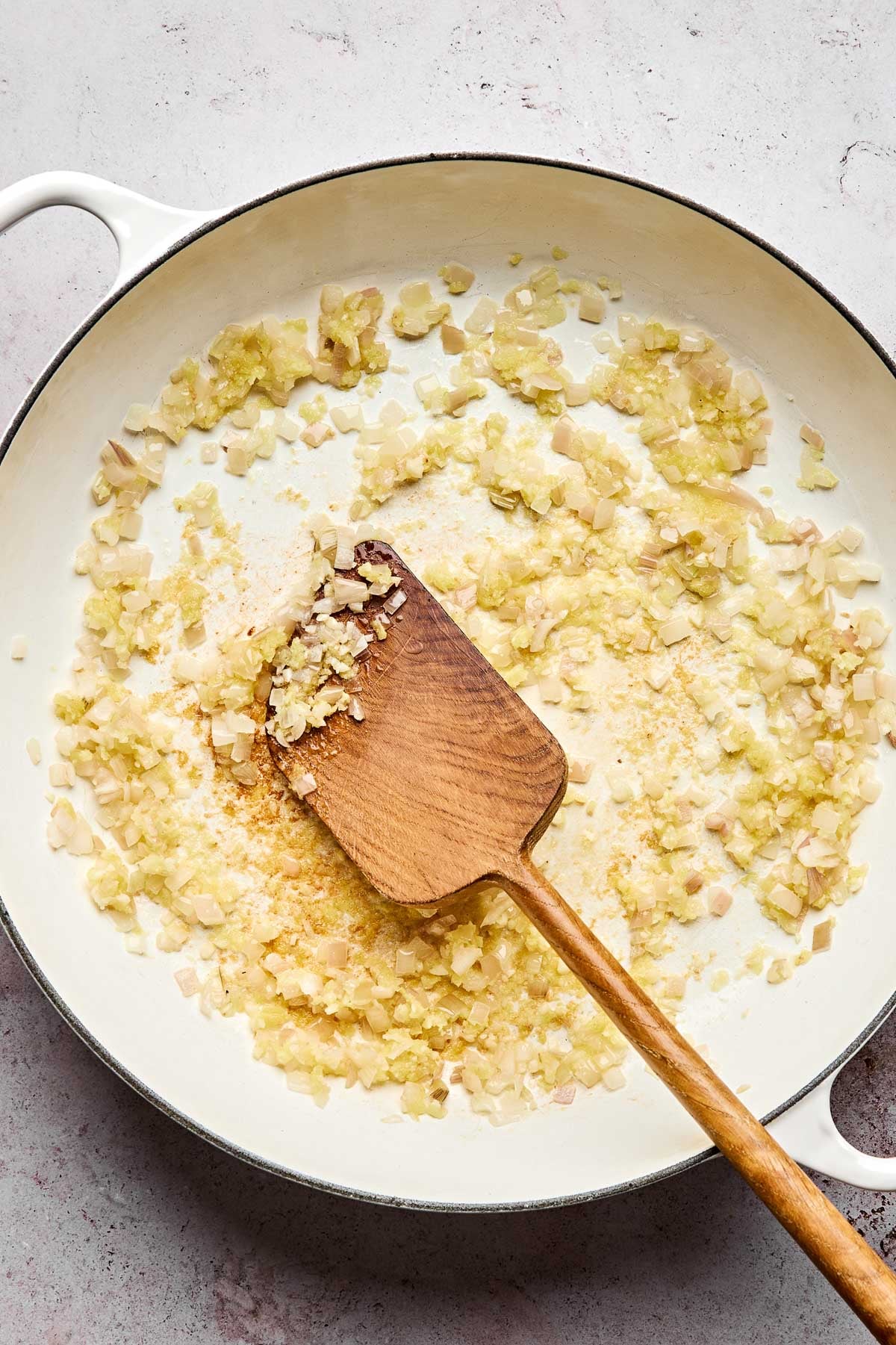 Chopped onions and garlic are being sautéed in a white skillet, stirred with a wooden spatula. The mixture appears to be softening and turning golden.