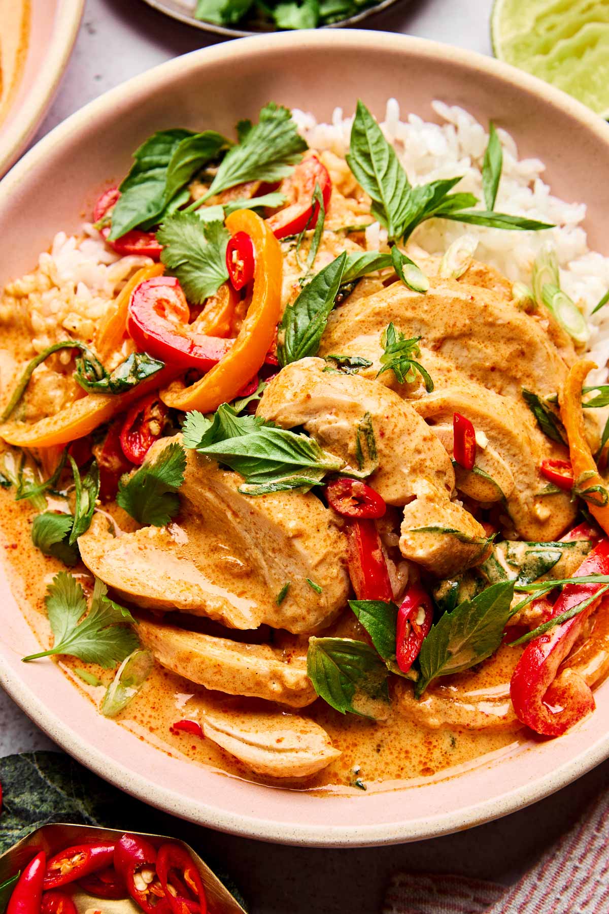 A bowl of Thai red curry with sliced chicken, bell peppers, red chili, fresh herbs, and white rice, served in a pink dish.