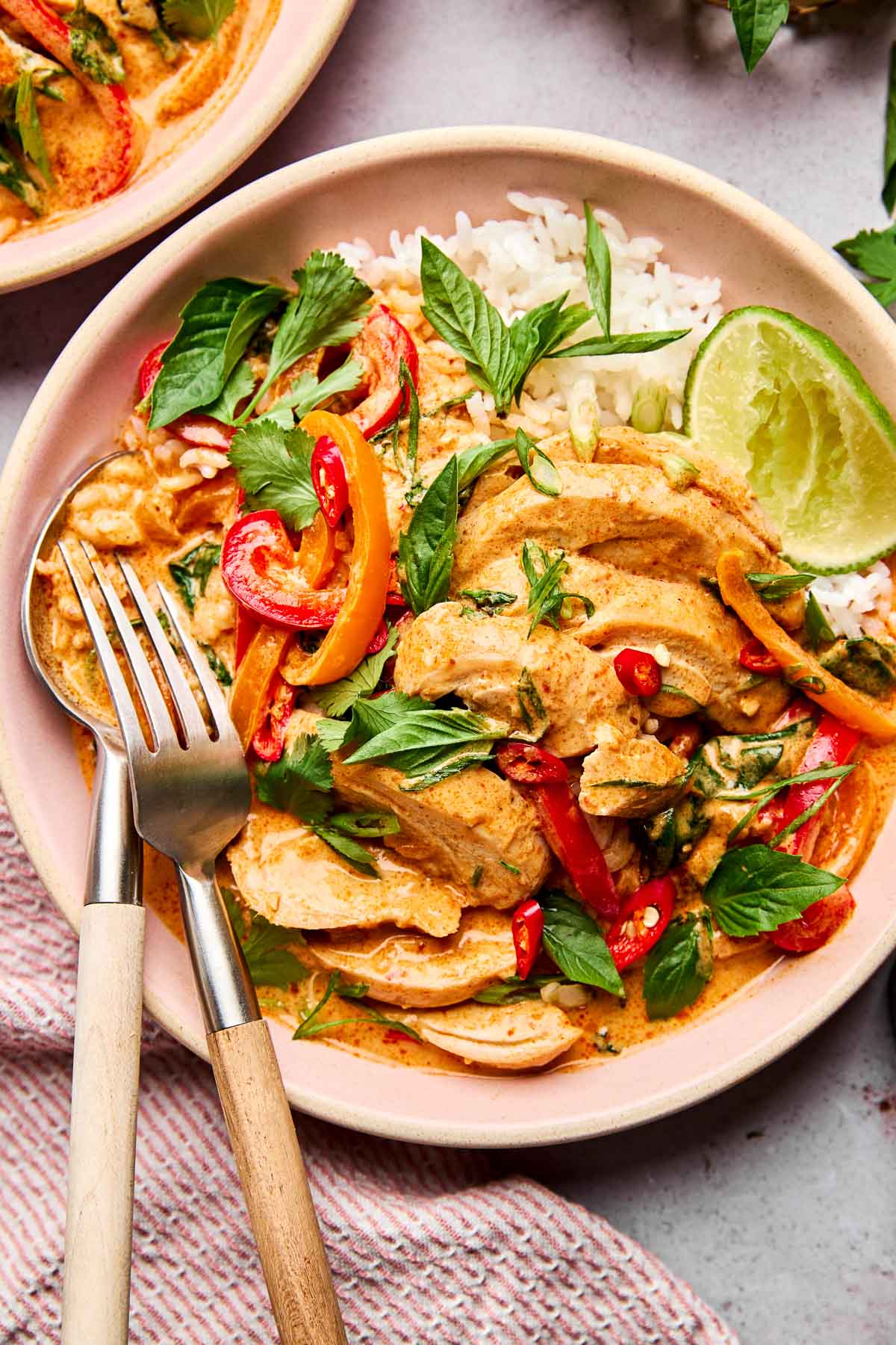 A bowl of Thai red curry with sliced chicken, bell peppers, fresh herbs, a lime wedge, and white rice, served with a fork and spoon on the side.
