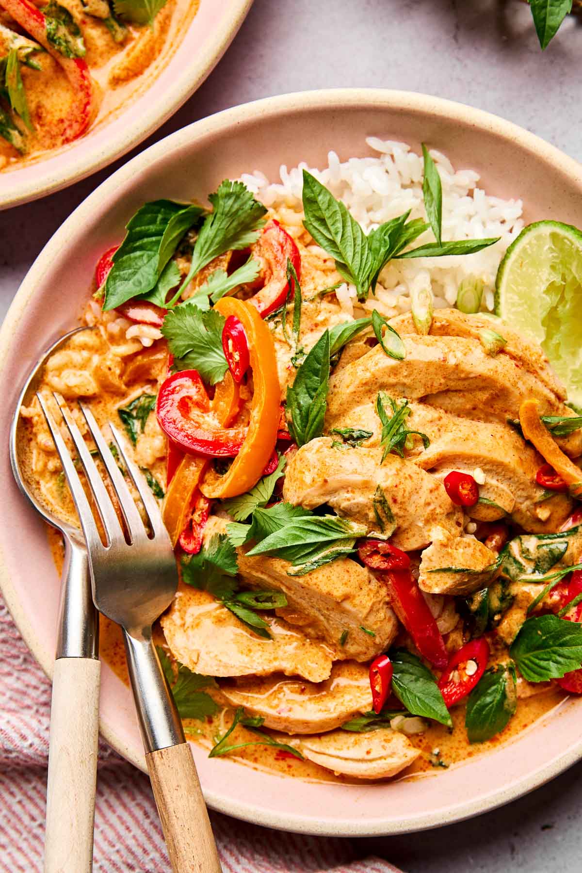A bowl of Thai red curry with sliced chicken, bell peppers, and fresh herbs, served with white rice and a lime wedge. A fork and spoon rest on the side of the bowl.