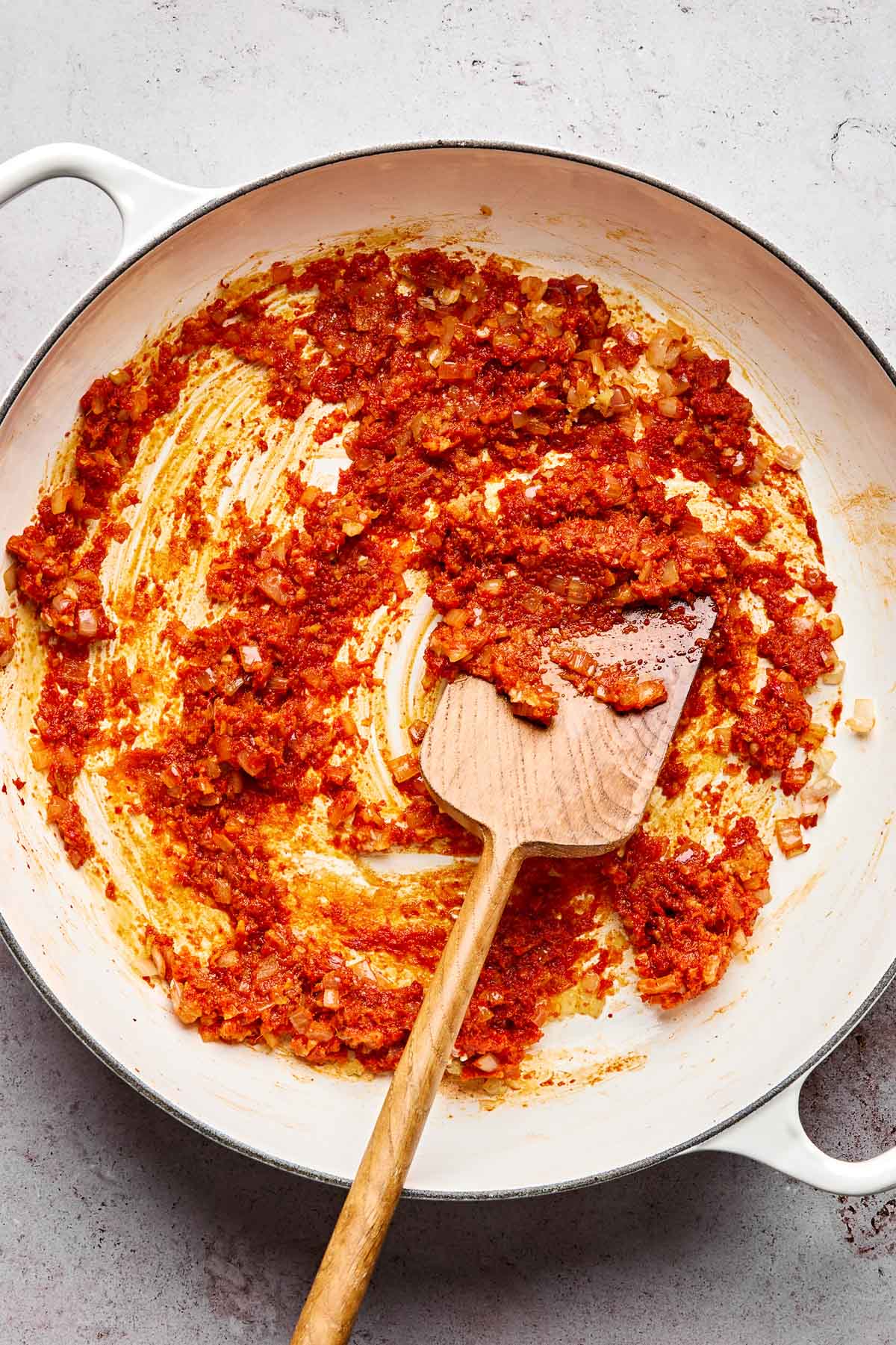 A white pan contains a mixture of tomato paste, chopped onions, and spices being sautéed, with a wooden spatula resting inside. The mixture is spread unevenly across the bottom of the pan.