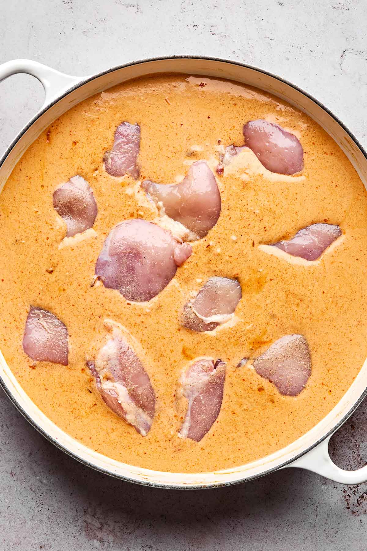 Raw pieces of chicken are submerged in an orange-colored creamy marinade inside a round white pot on a light gray surface.