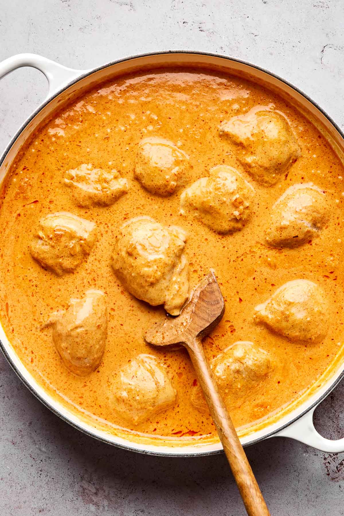 A large white pot filled with creamy orange butter chicken curry, with several pieces of chicken in the sauce. A wooden spoon rests in the pot, stirring the curry. The background is a light stone surface.