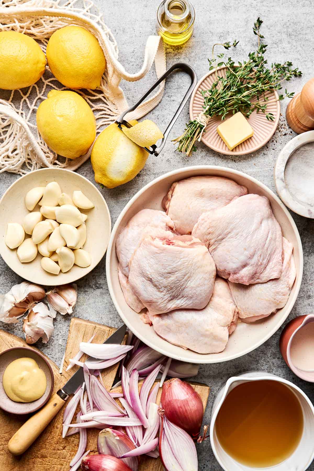 Raw chicken thighs on a plate surrounded by lemon, garlic cloves, shallots, fresh thyme, butter, olive oil, Dijon mustard, salt, pepper, and a bowl of broth on a countertop.