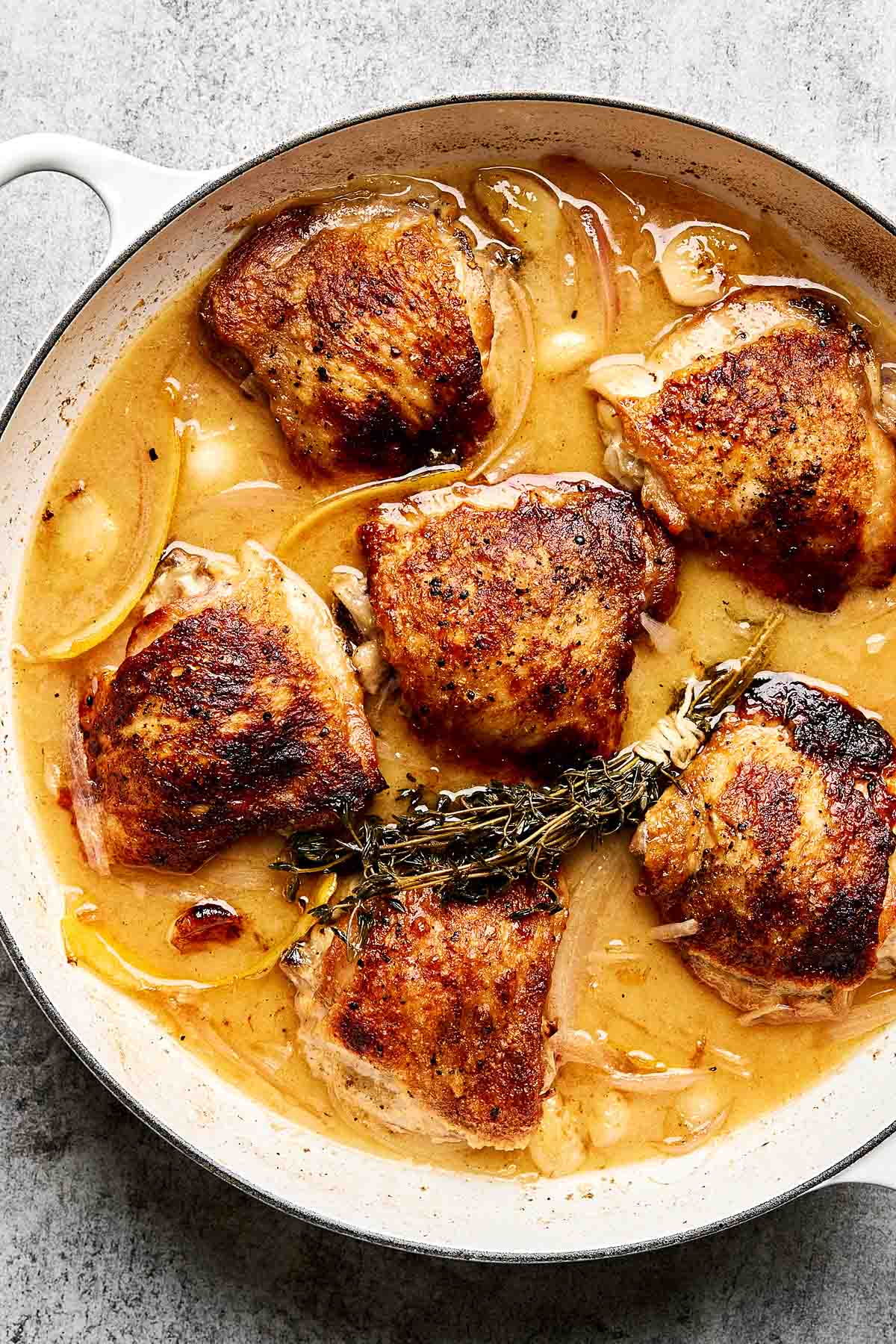 Golden-brown chicken thighs simmer in a creamy sauce with sliced onions, garlic, and sprigs of fresh thyme in a white skillet on a gray surface.