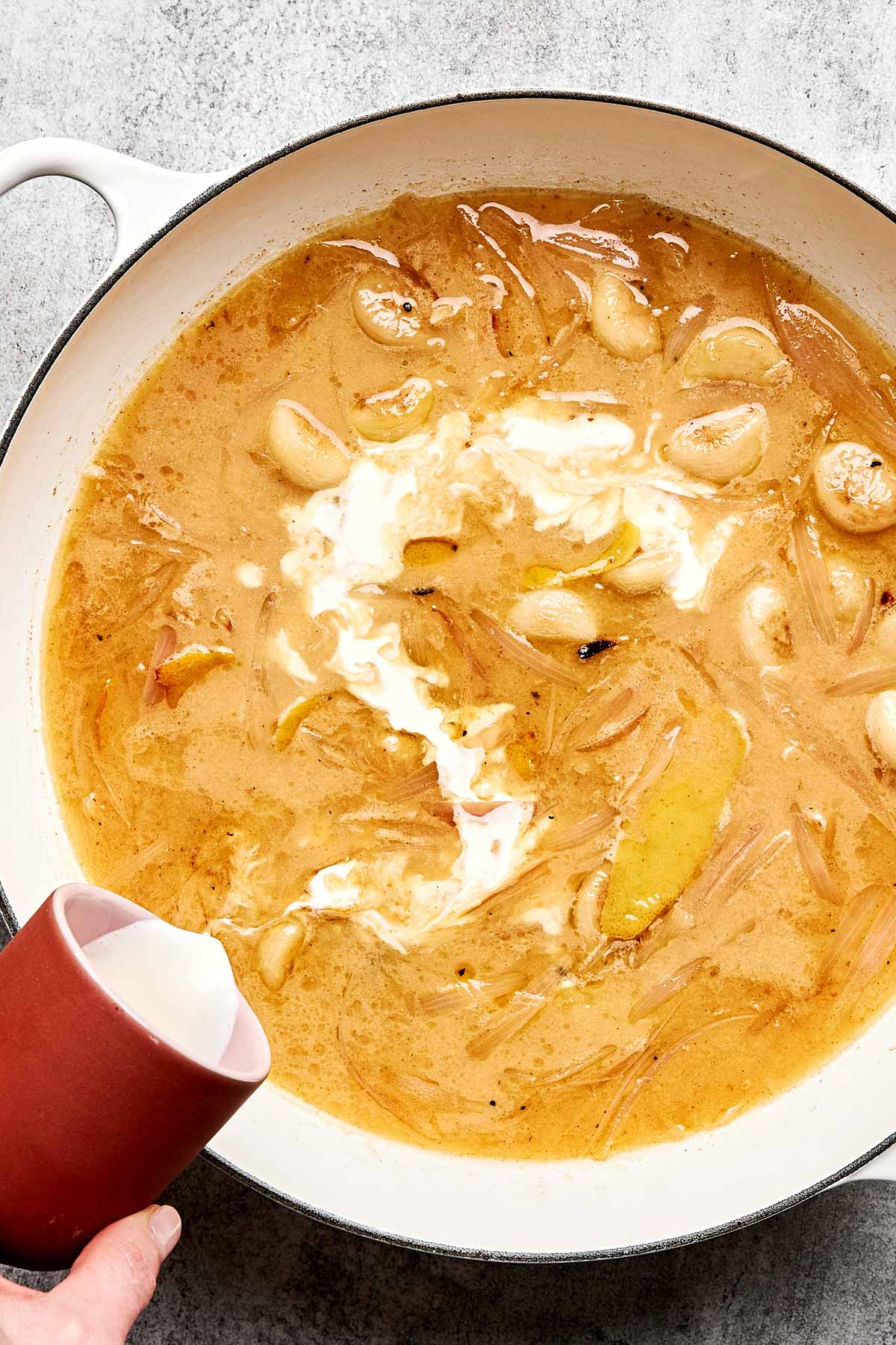 A hand pours cream from a red cup into a pot of golden brown soup with sliced onions and whole garlic cloves, creating a swirling effect on the surface.