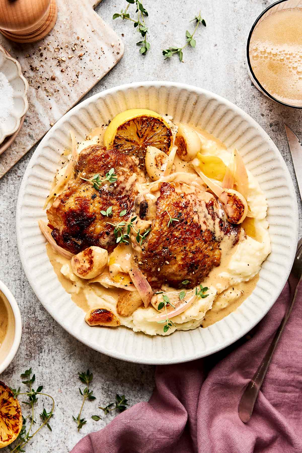 A plate of roasted chicken thighs served on creamy mashed potatoes, garnished with garlic cloves, herbs, and grilled lemon slices, with a light brown sauce. A drink, napkin, and herbs are nearby.