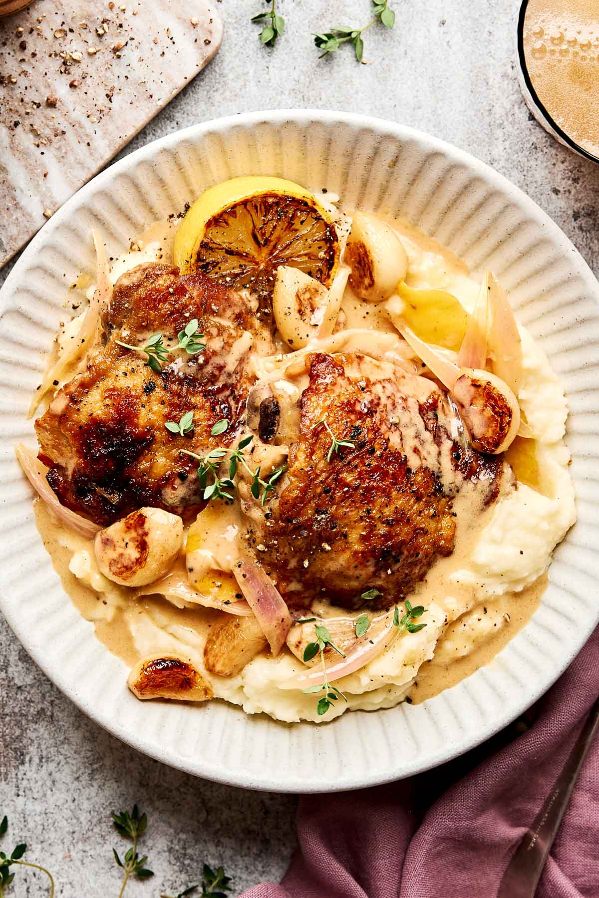 A plate of creamy mashed potatoes topped with two pieces of roasted chicken, caramelized garlic cloves, onion slices, lemon, fresh herbs, and a creamy sauce.