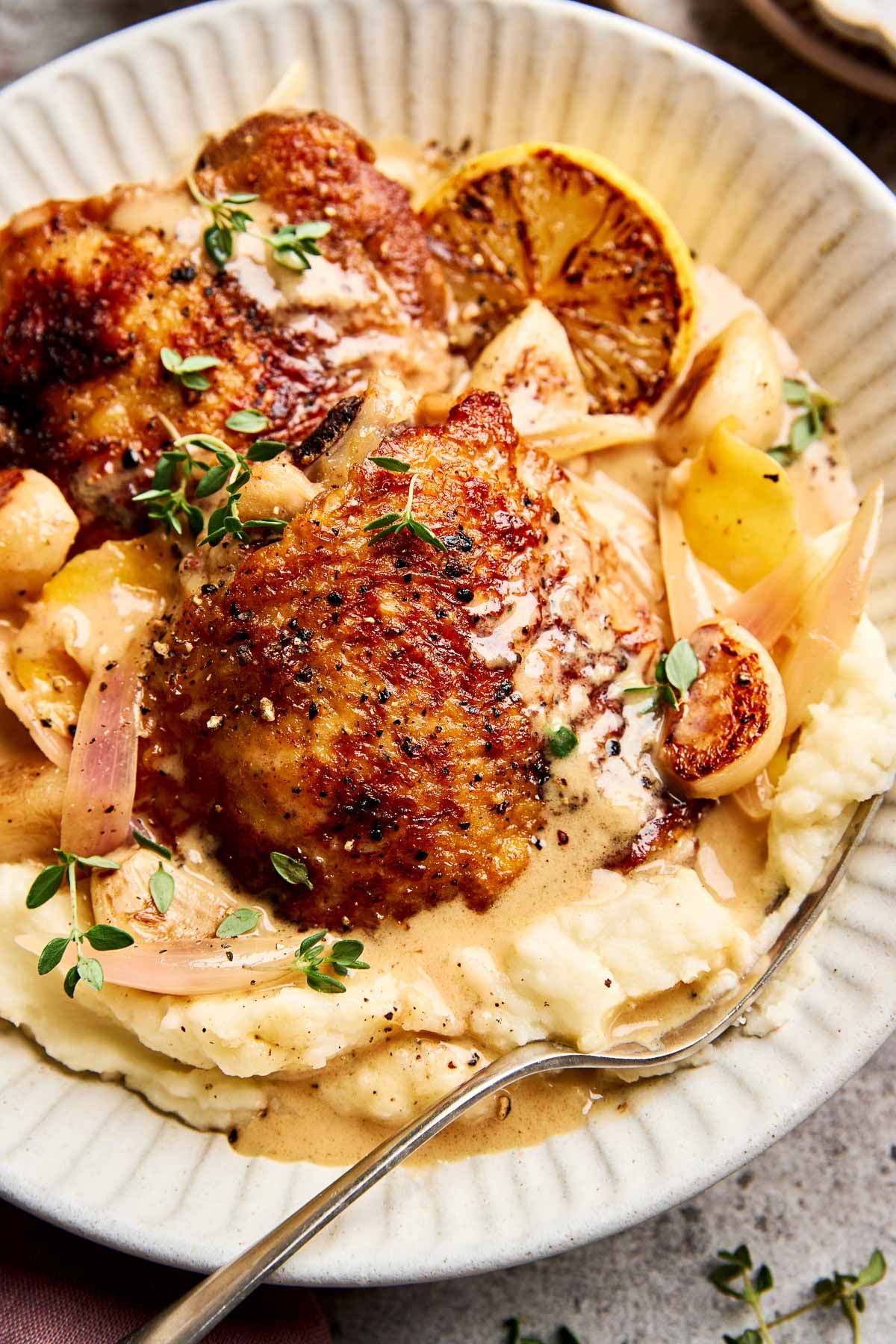 A plate of creamy mashed potatoes topped with golden-brown roasted chicken thighs, lemon slices, shallots, herbs, and a creamy gravy, served with a fork.