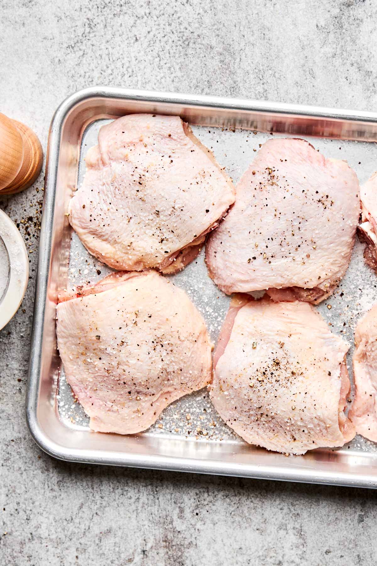 Raw chicken thighs with skin are arranged on a metal baking tray, seasoned with salt and black pepper. The tray rests on a light gray countertop, with a wooden pepper grinder and a white plate nearby.