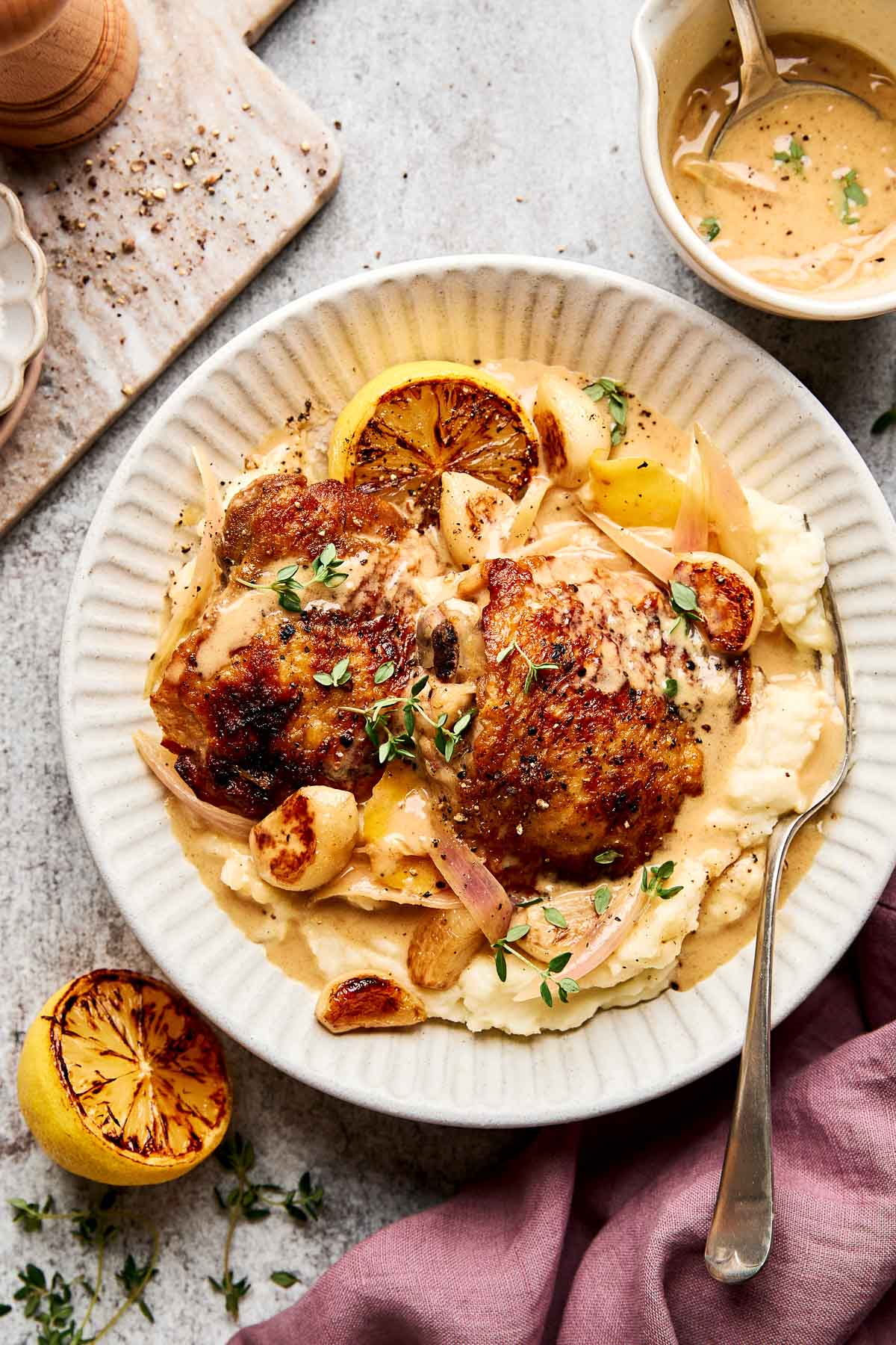 A plate of creamy mashed potatoes topped with roasted chicken, caramelized garlic, herbs, and a grilled lemon slice, served with a side of sauce and garnished with fresh thyme. A fork rests on the plate.