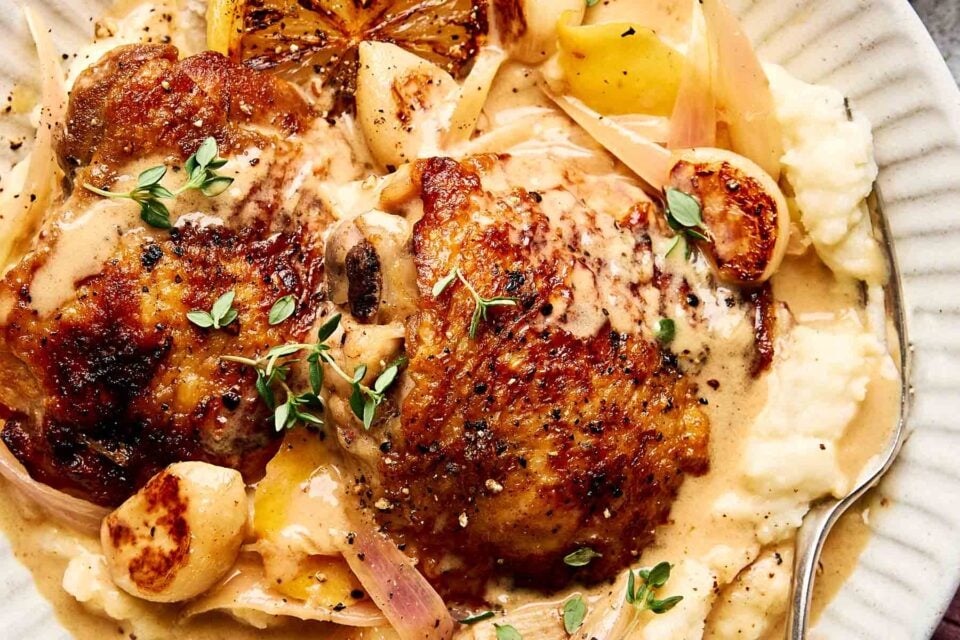 A plate of creamy mashed potatoes topped with crispy, golden-brown chicken thighs, roasted garlic, shallots, lemon, fresh thyme, and a rich sauce. A spoon rests on the side of the dish.