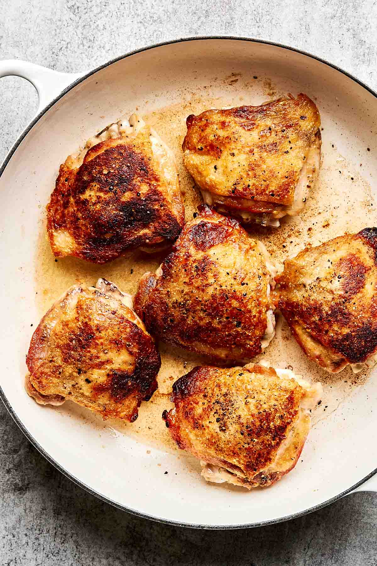 Six golden-brown, crispy chicken thighs are cooking in a white skillet on a gray surface, with visible seasoning and rendered juices in the pan.