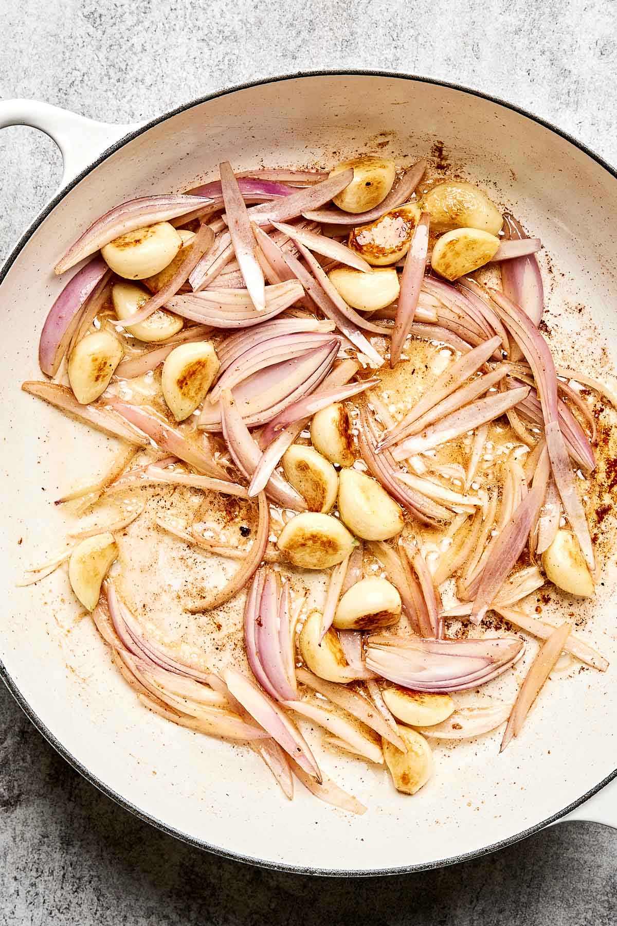 Thinly sliced red onions and whole garlic cloves sauté in oil in a white skillet on a gray surface. The ingredients are lightly browned and spread across the pan.
