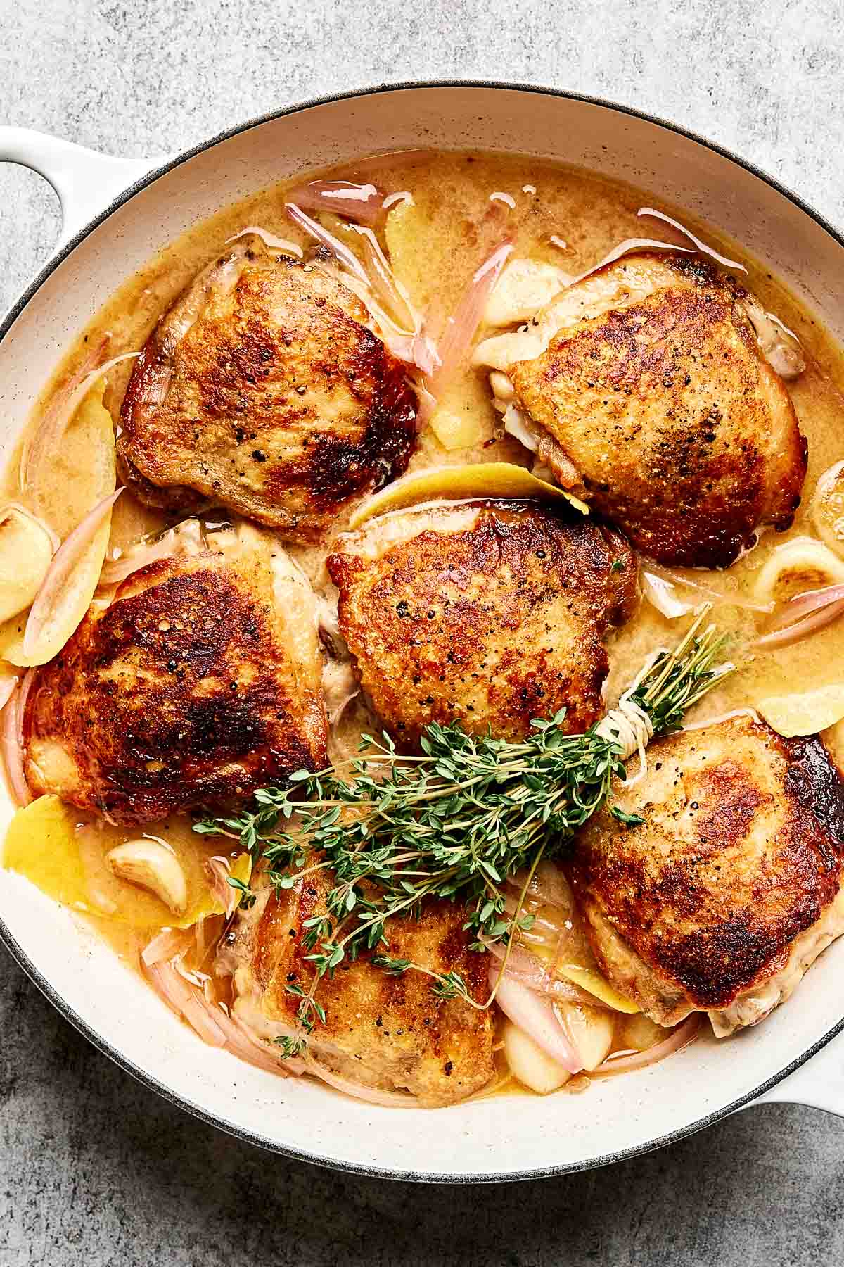 A skillet filled with six golden-brown, crispy chicken thighs simmering in a light sauce with sliced onions and apples, garnished with fresh thyme sprigs.