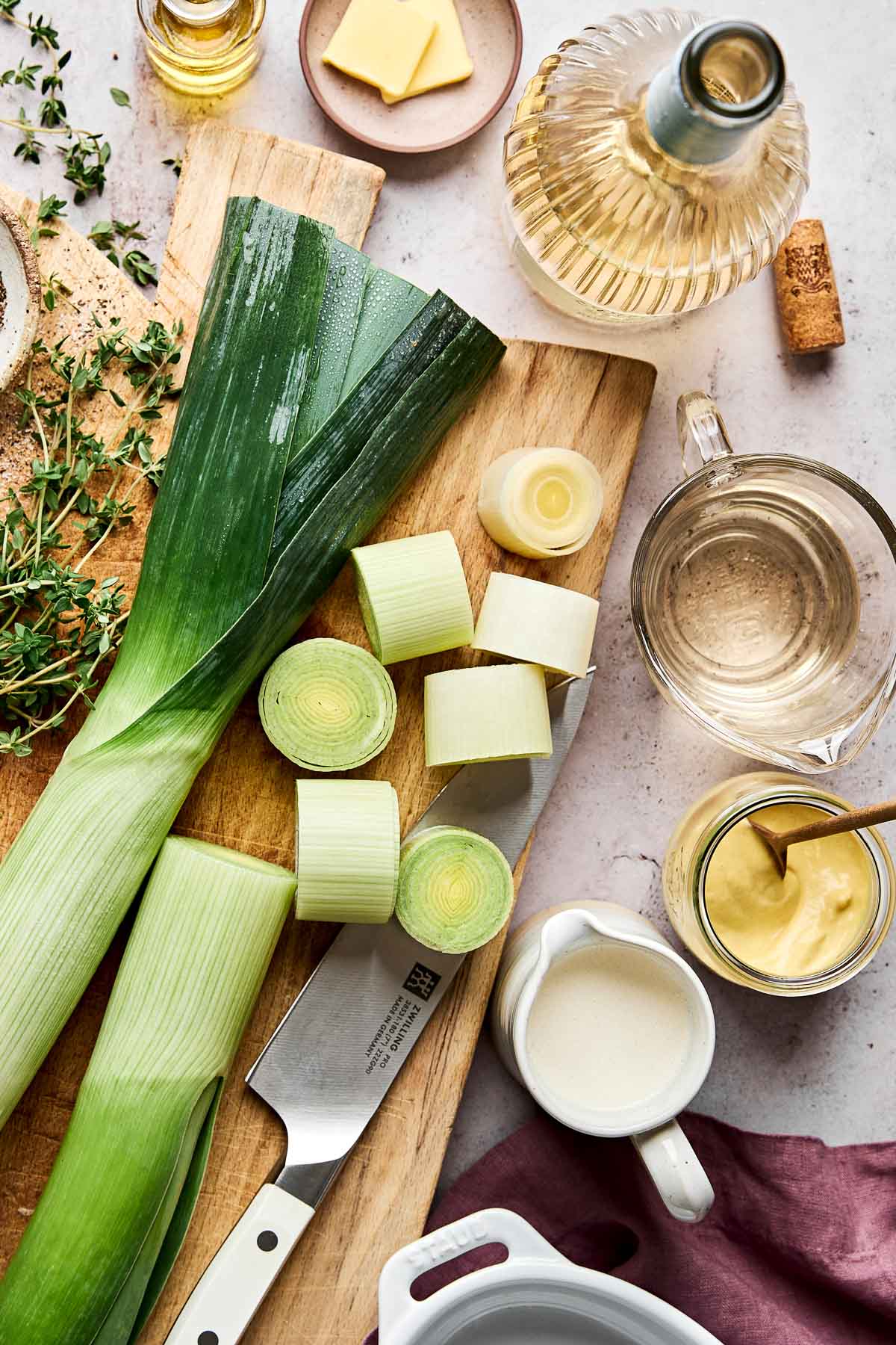 A wooden cutting board with sliced leeks, a knife, fresh herbs, butter, white wine, cream, Dijon mustard, and a measuring cup with broth, arranged for cooking.
