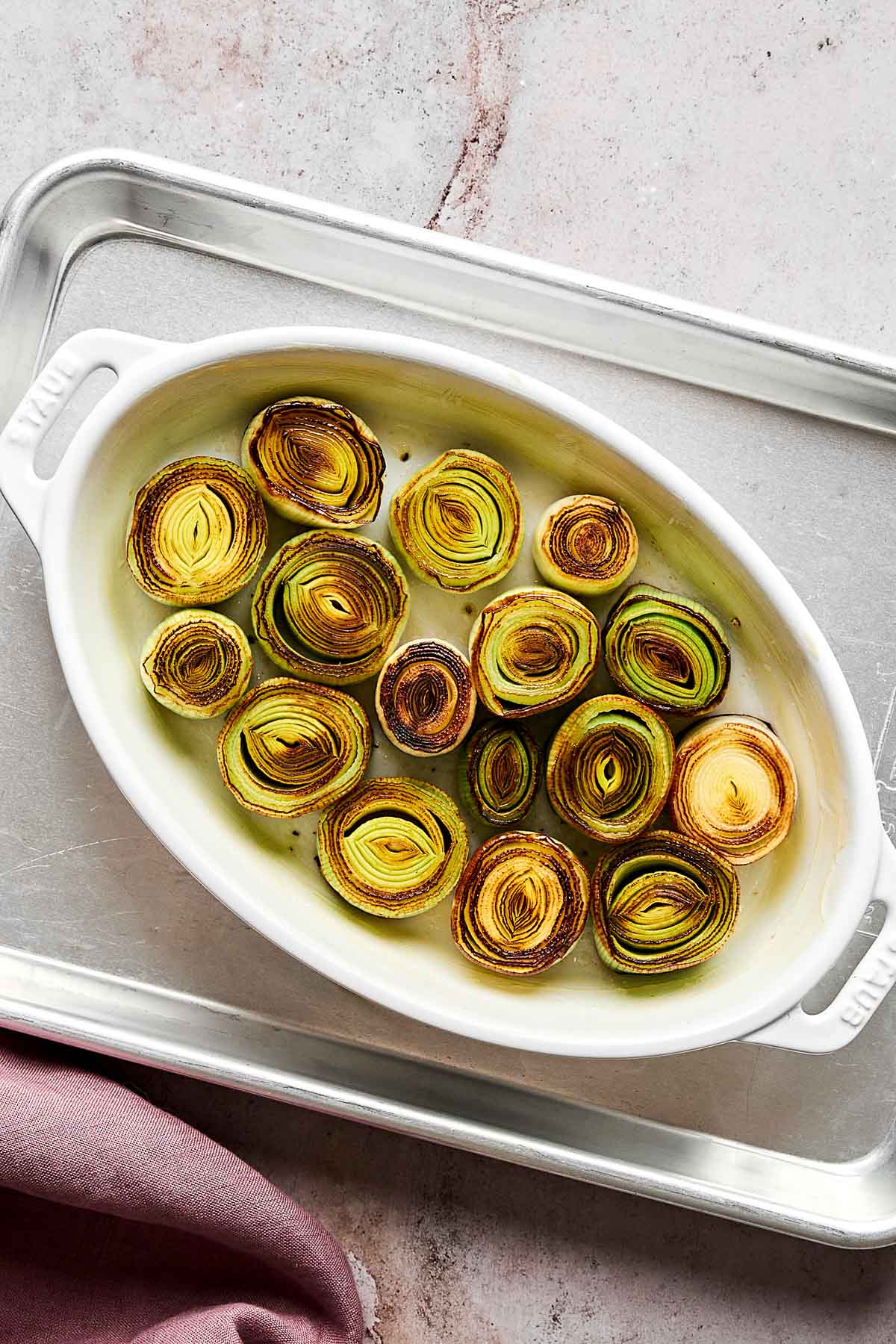 Oval white baking dish with roasted leek rounds arranged in a single layer, showing golden-brown and green edges. The dish sits on a metal tray atop a light, textured surface, with a folded mauve cloth nearby.