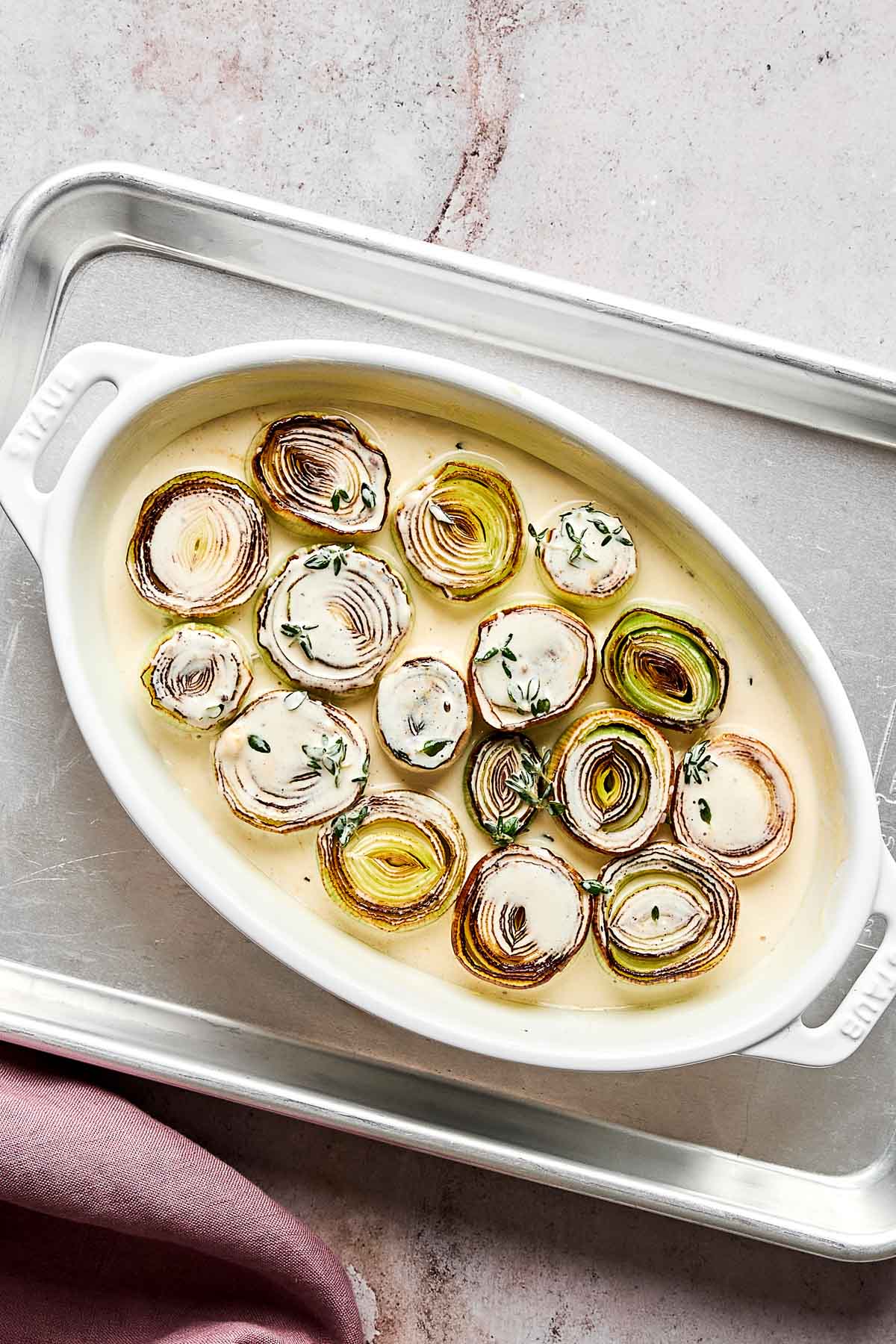An oval white baking dish filled with sliced leeks arranged in a creamy sauce, garnished with fresh thyme sprigs, sits on a metal tray with a mauve cloth nearby.
