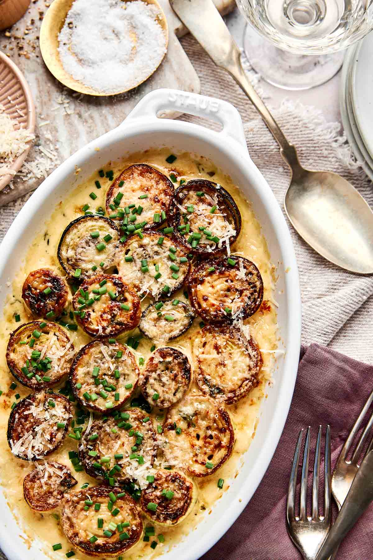 A white oval dish filled with baked zucchini rounds topped with grated cheese and chopped chives, set on a table with utensils, plates, and a glass of water nearby.