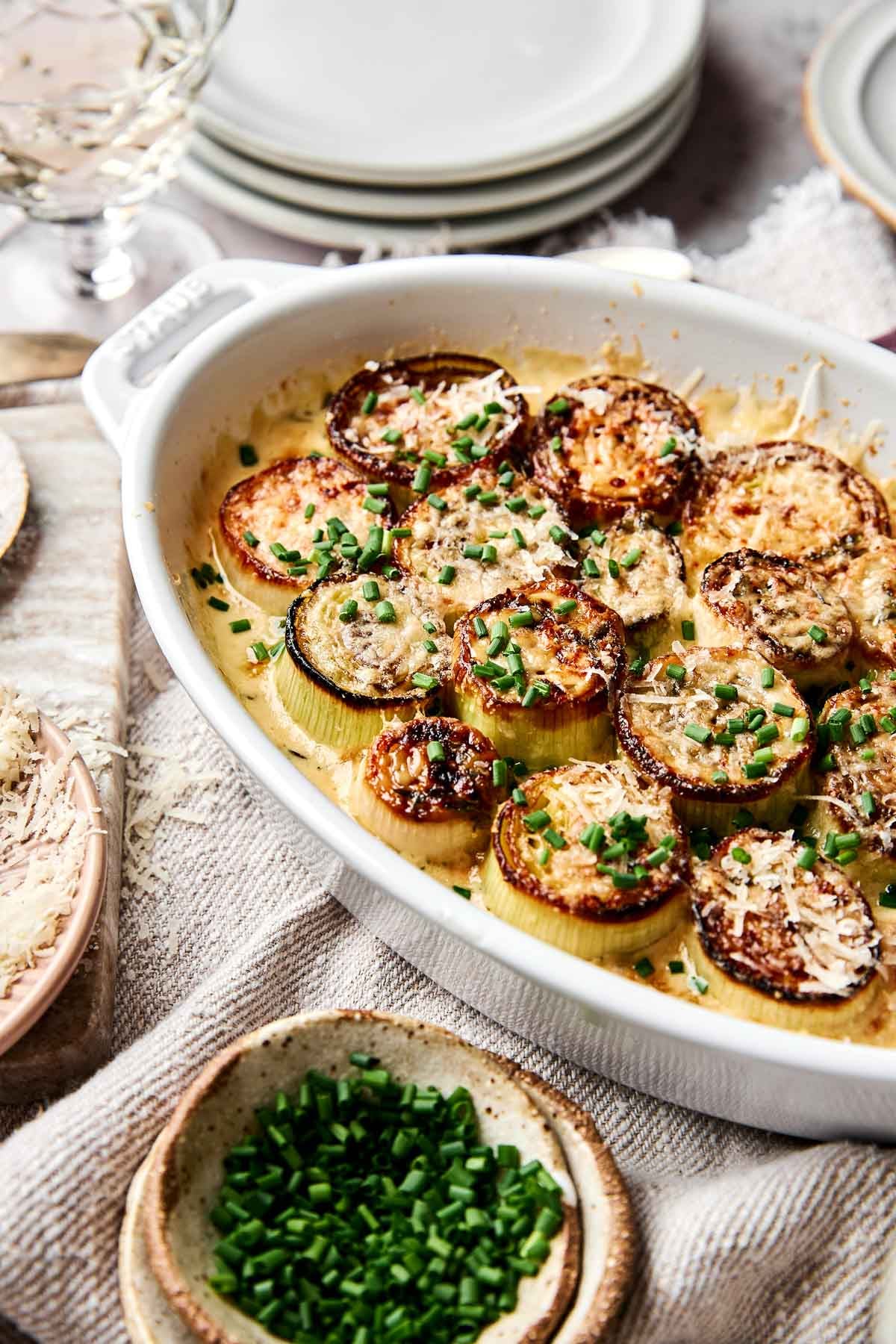 A white baking dish filled with creamy, golden-brown roasted leeks topped with grated cheese and chopped chives, set on a textured cloth with plates and a bowl of fresh chives nearby.