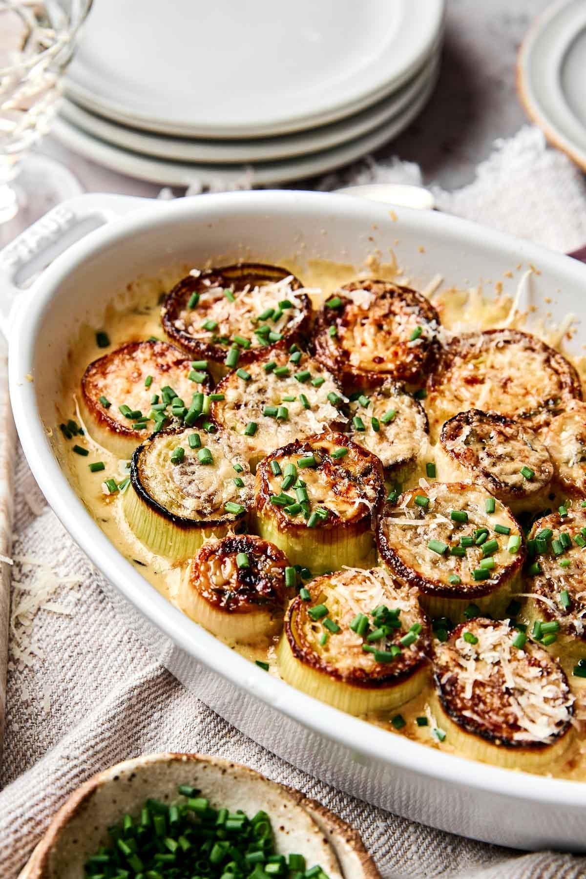 A white baking dish filled with browned roasted leek rounds topped with grated cheese and chopped chives, set on a textured cloth with stacked plates and a bowl of chives nearby.