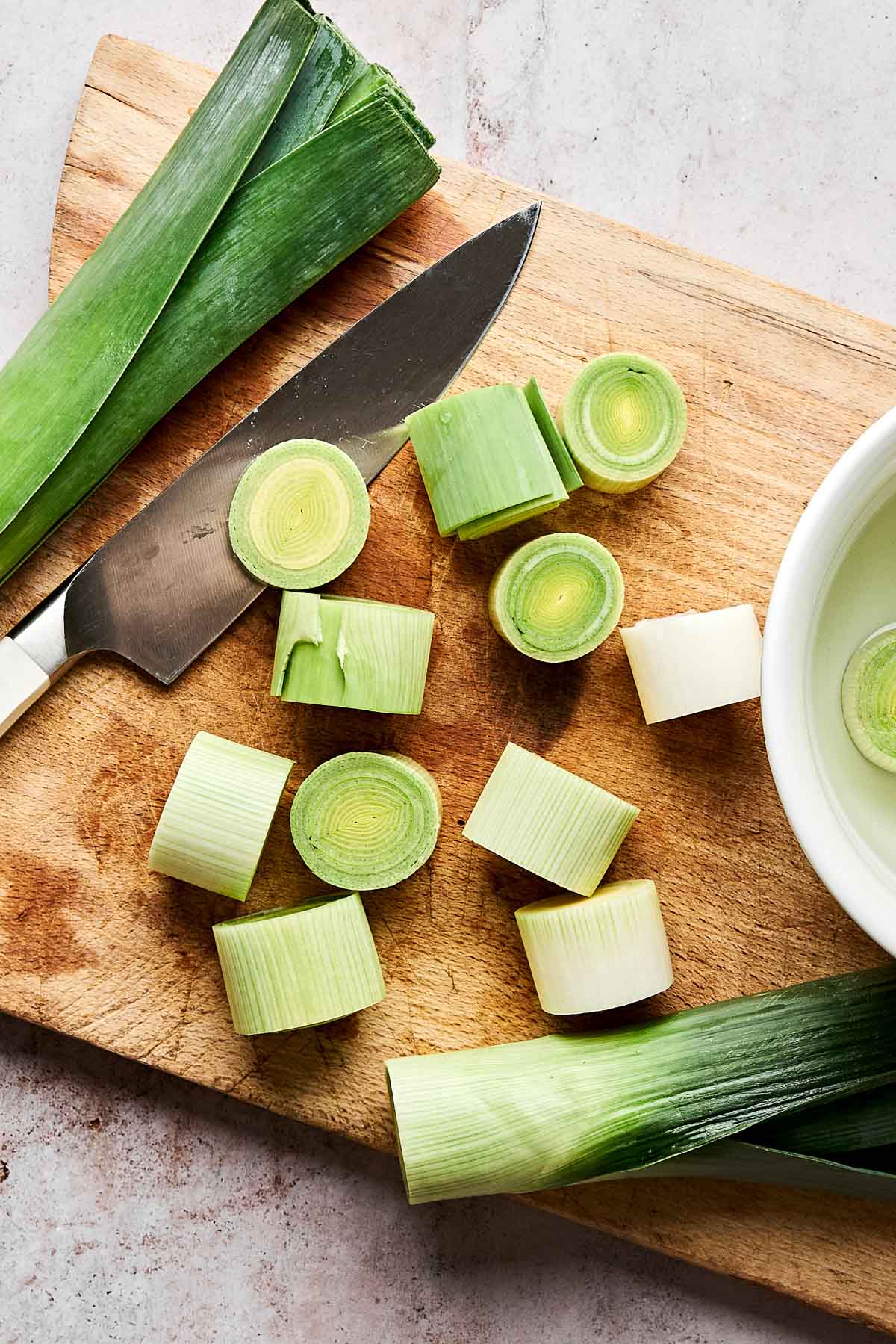 Sliced leeks on a wooden cutting board with a knife and whole leek leaves, next to a bowl of water on a light countertop.