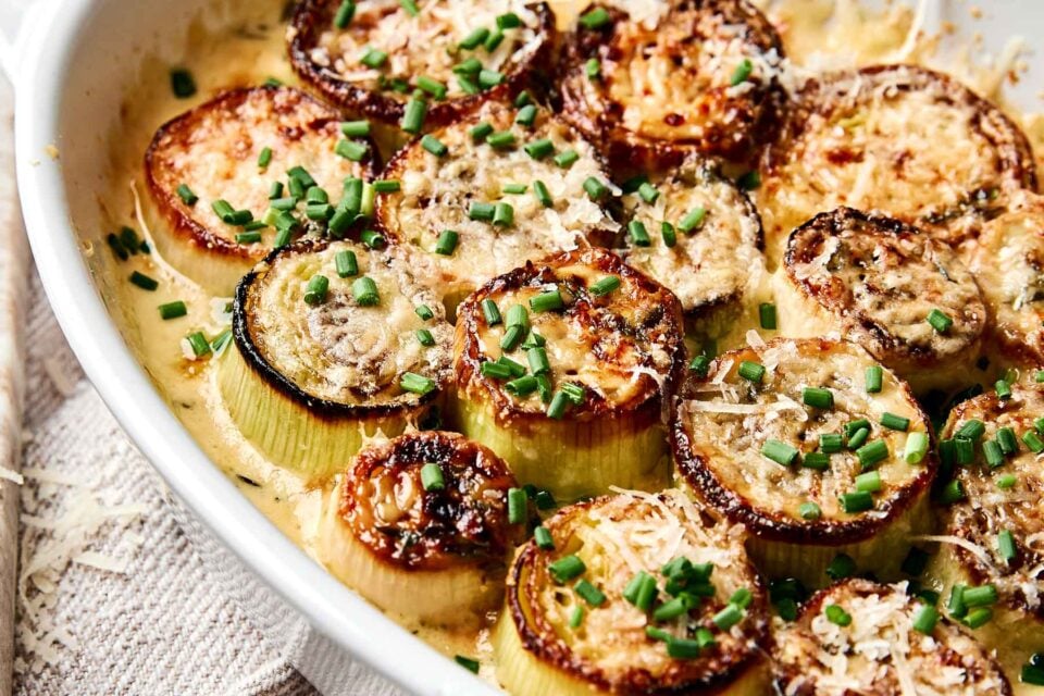 A close-up of roasted leeks topped with melted cheese and chopped chives in a creamy sauce, served in a white baking dish.