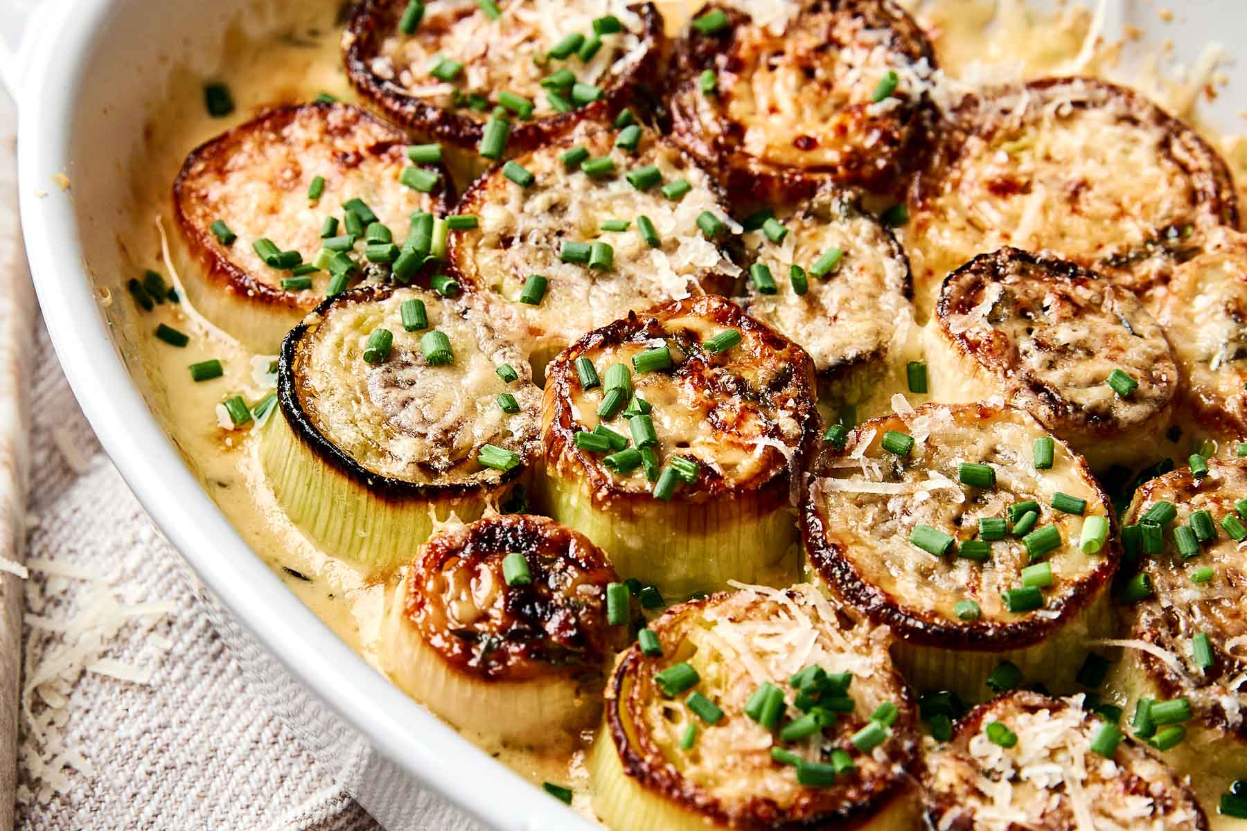 A close-up of roasted leeks topped with melted cheese and chopped chives in a creamy sauce, served in a white baking dish.