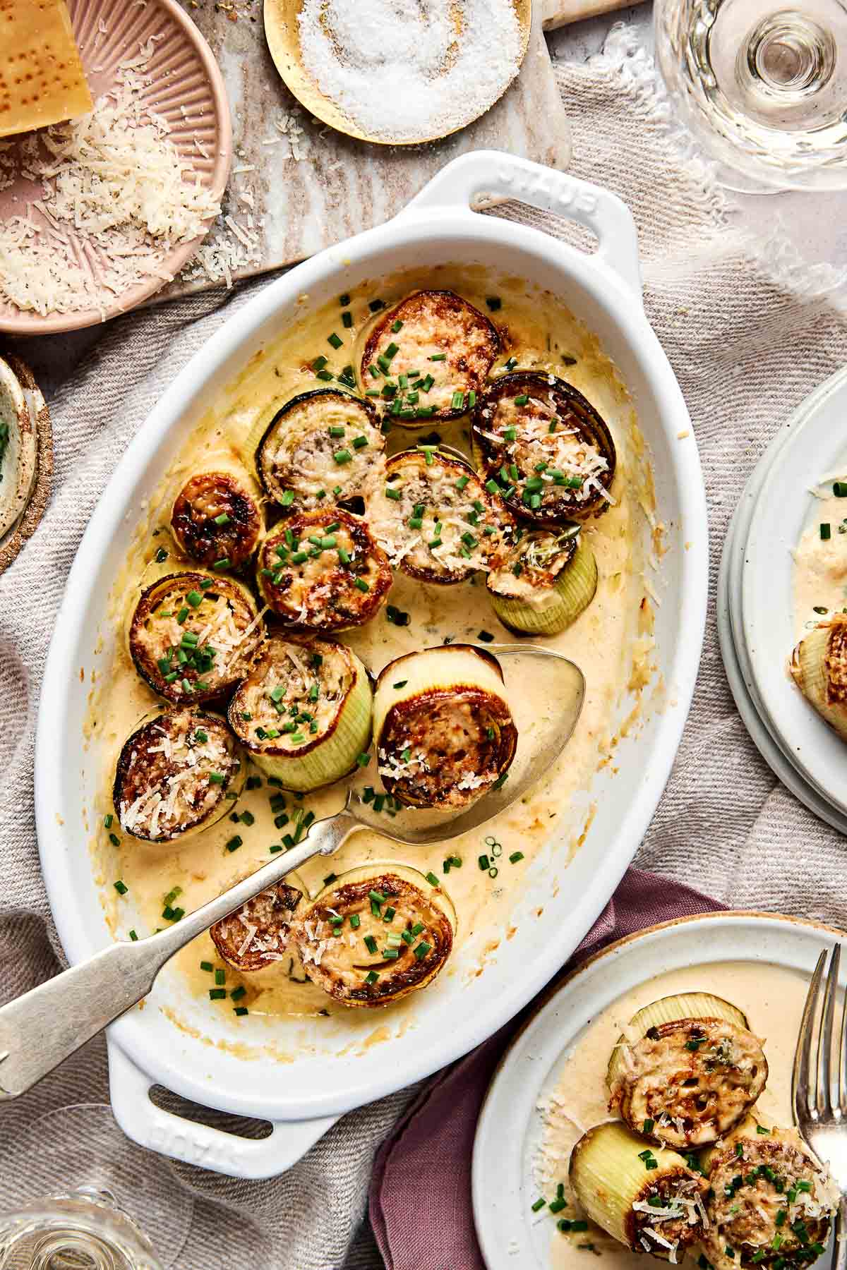 A white oval baking dish filled with baked, sliced leeks topped with a creamy sauce, grated cheese, and chopped chives. A large serving spoon rests in the dish, with plates and glasses nearby on a set table.