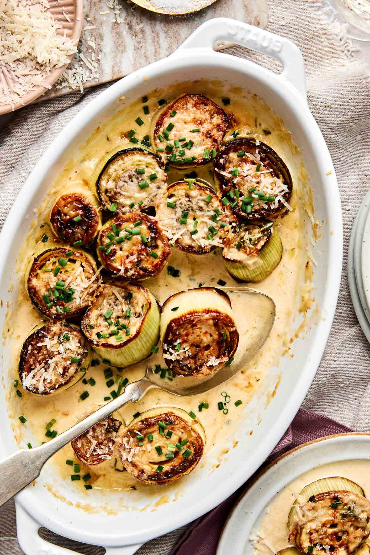 Sliced baked leeks topped with grated cheese and chopped chives in a creamy sauce, served in a white oval baking dish with a serving spoon. Plates and extra cheese are nearby.