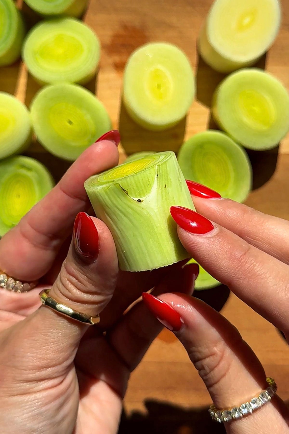 A person with red manicured nails holds a piece of sliced leek, with several more leek slices arranged on a wooden surface in the background.