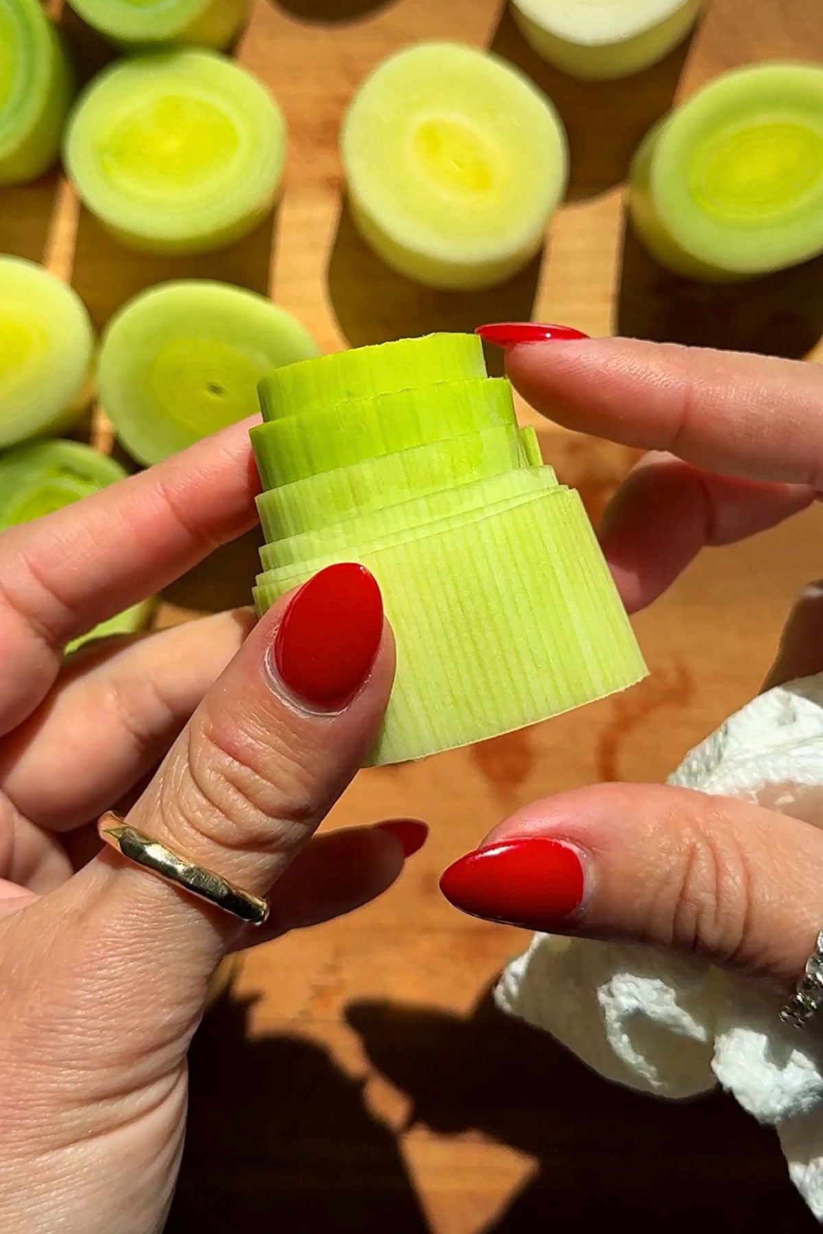 A person with bright red nail polish holds a cut piece of leek, showing its light green and white layers. More slices of leek are visible in the background on a wooden surface.