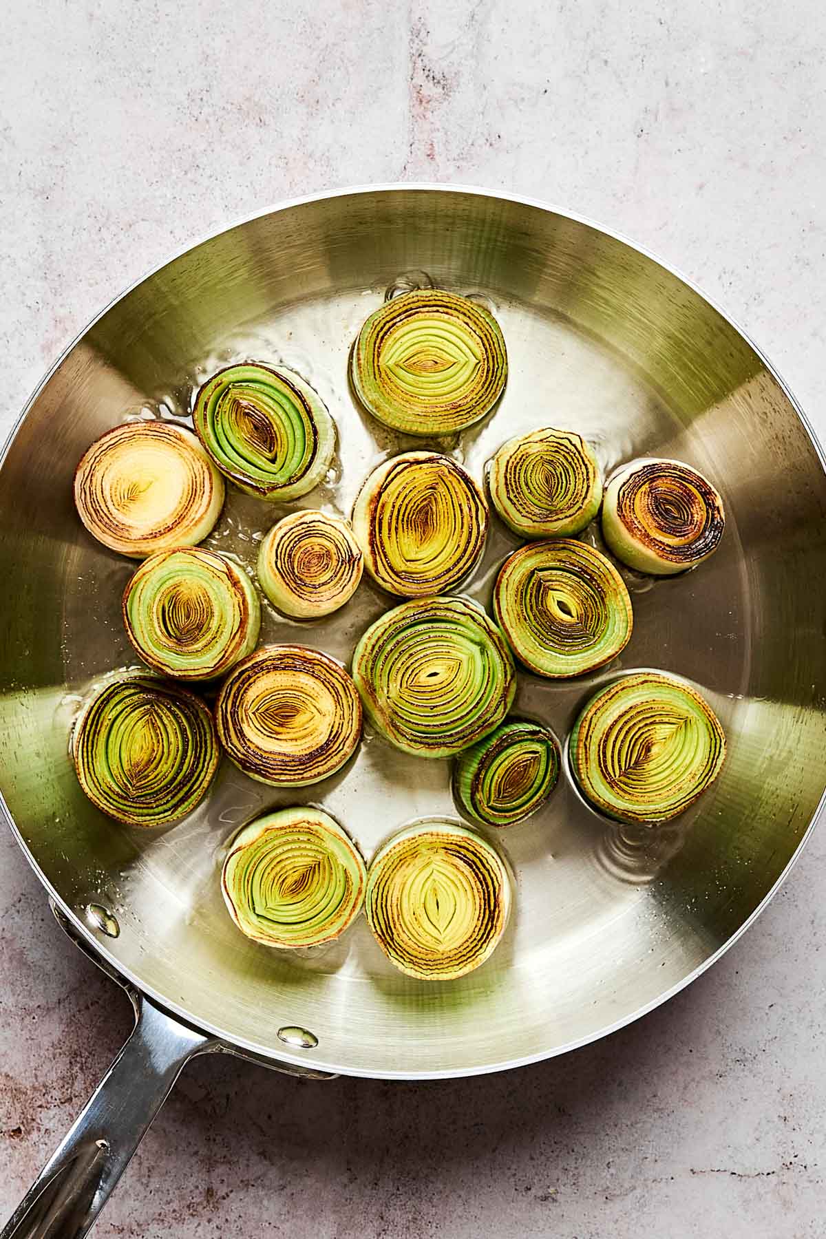 A stainless steel pan holds several round slices of leeks being sautéed, some with lightly browned edges, on a light-colored surface.