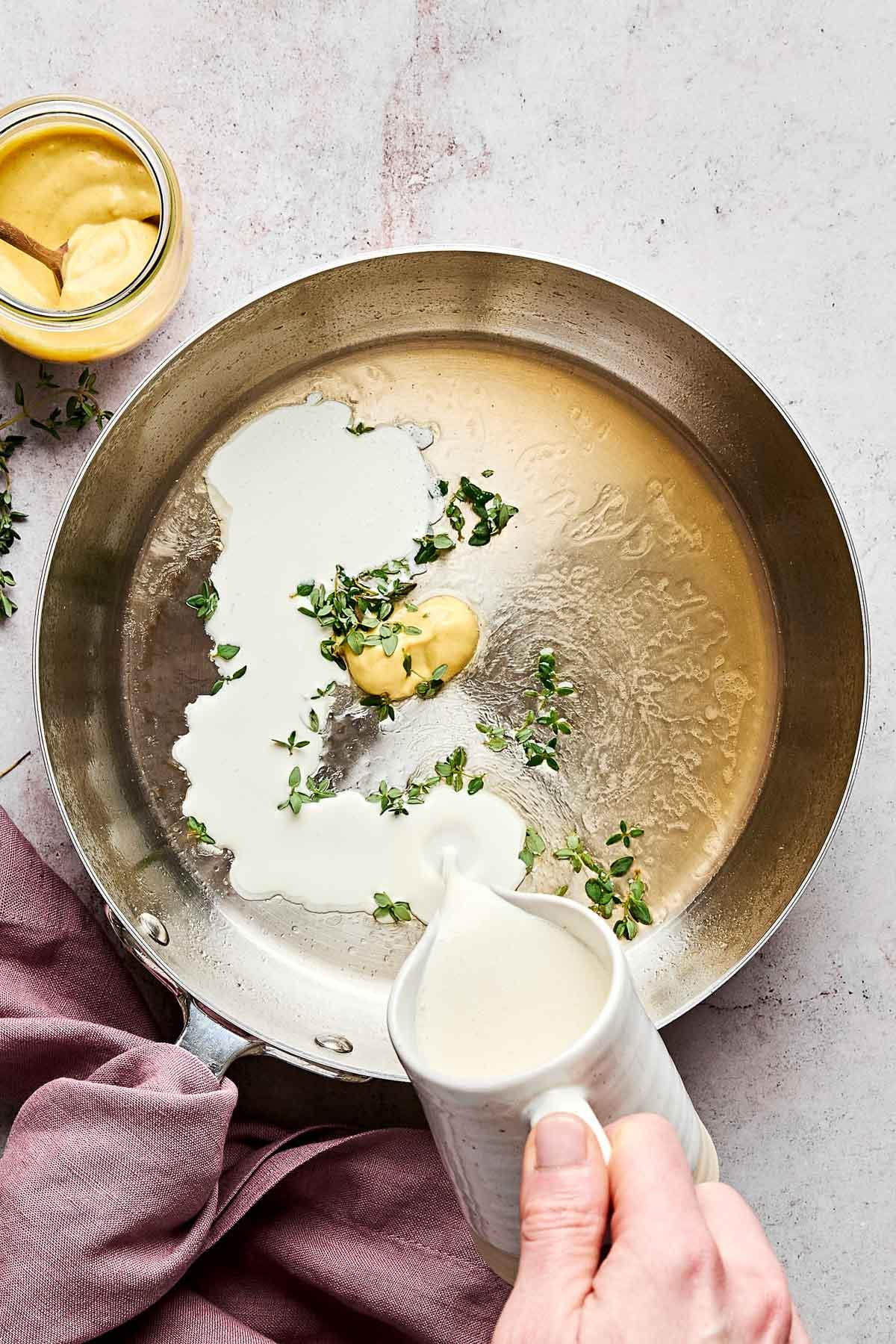 A hand pours cream from a small pitcher into a skillet with mustard, fresh thyme, and melted butter. A jar of mustard and a mauve cloth sit nearby on a light countertop.