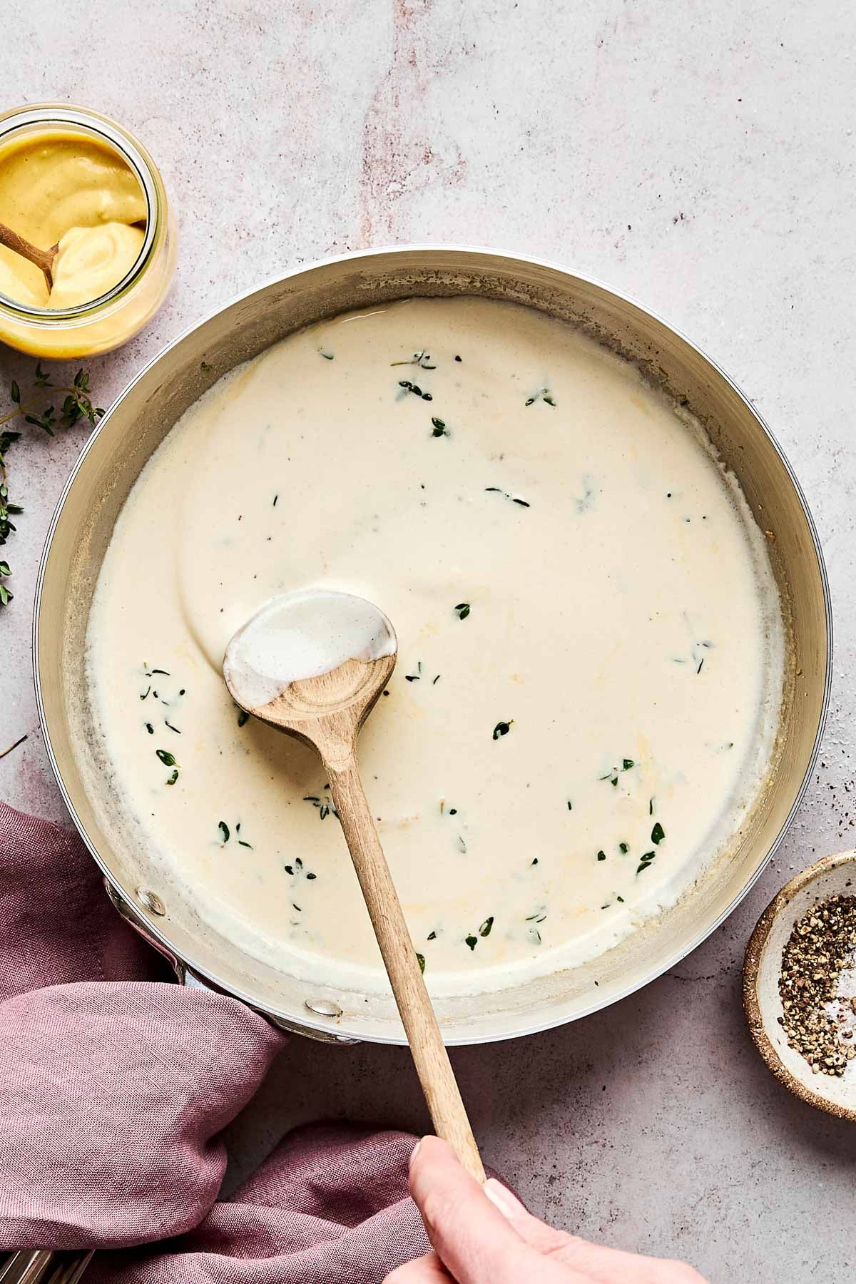 A hand stirs creamy white sauce with herbs in a pan using a wooden spoon; a jar of mustard, pink cloth, and bowl of pepper are nearby on a light countertop.