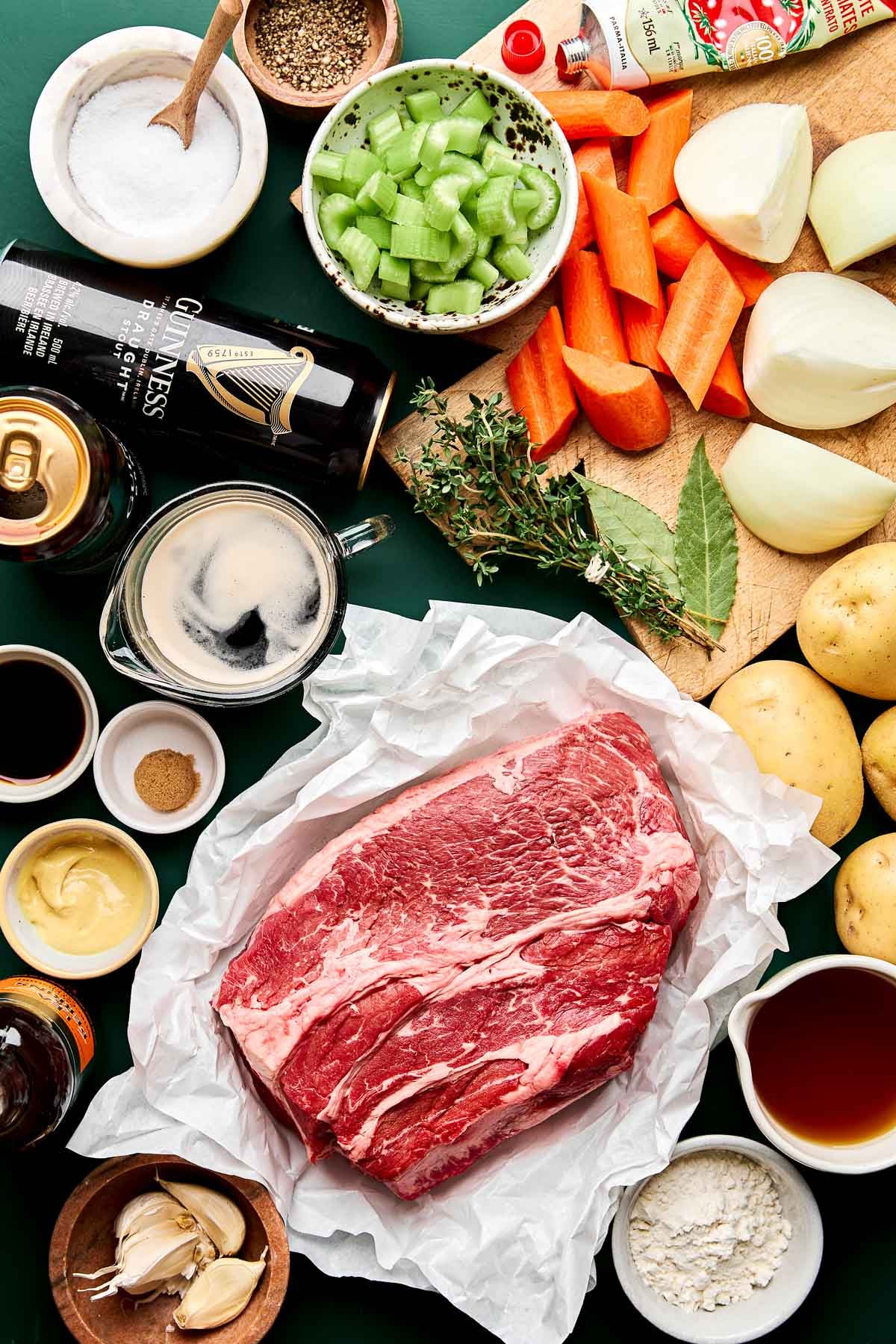 Ingredients for Guinness Pot Roast A flat lay of beef chuck roast, carrots, celery, onions, potatoes, garlic, mustard, seasonings, herbs, beer, Worcestershire sauce, beef broth, tomato paste, and flour on a green surface, ready for cooking.