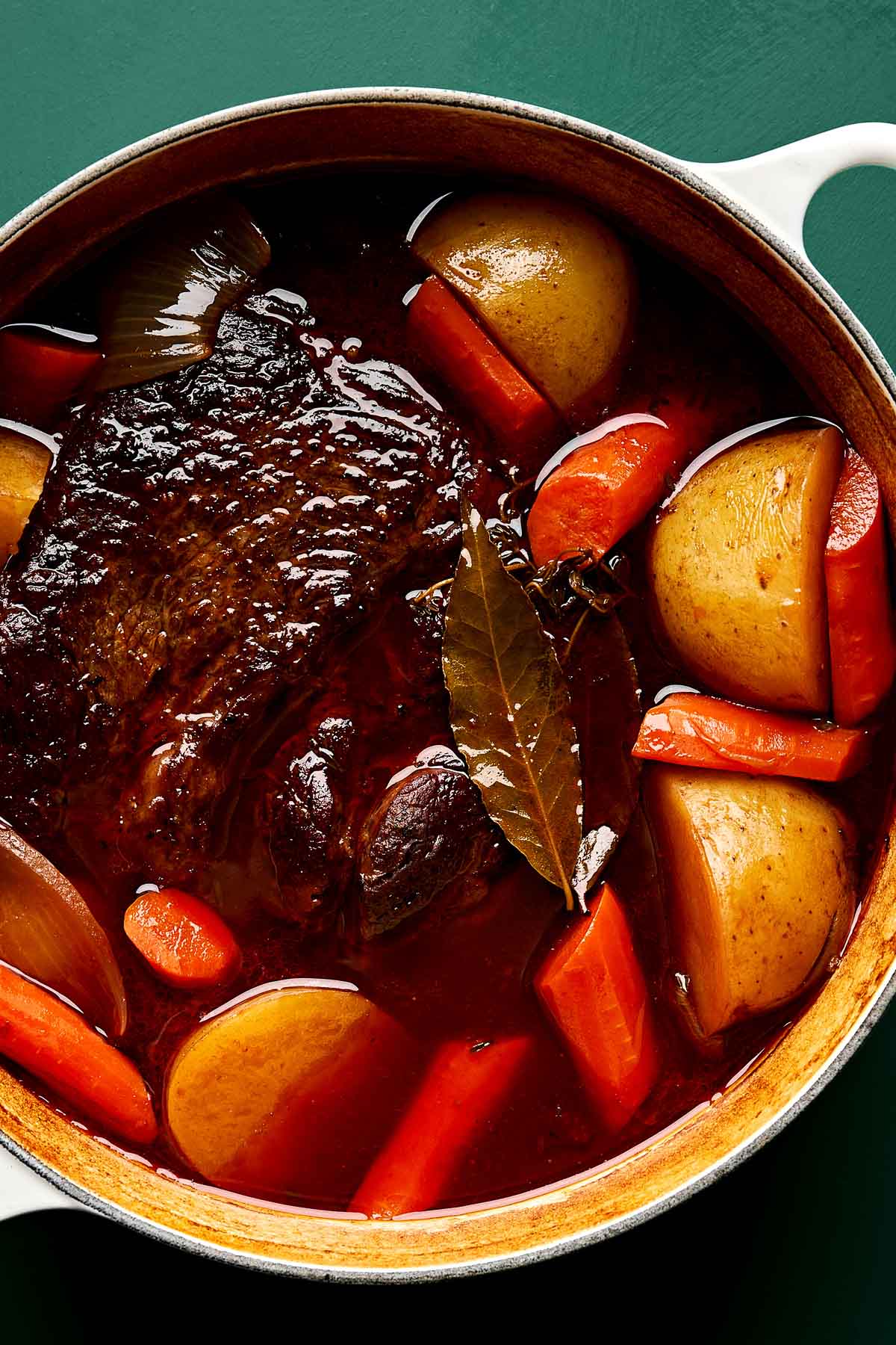 Braising Pot Roast A pot roast with beef, potatoes, carrots, onions, and bay leaves, all simmering in a rich, brown broth inside a white Dutch oven.