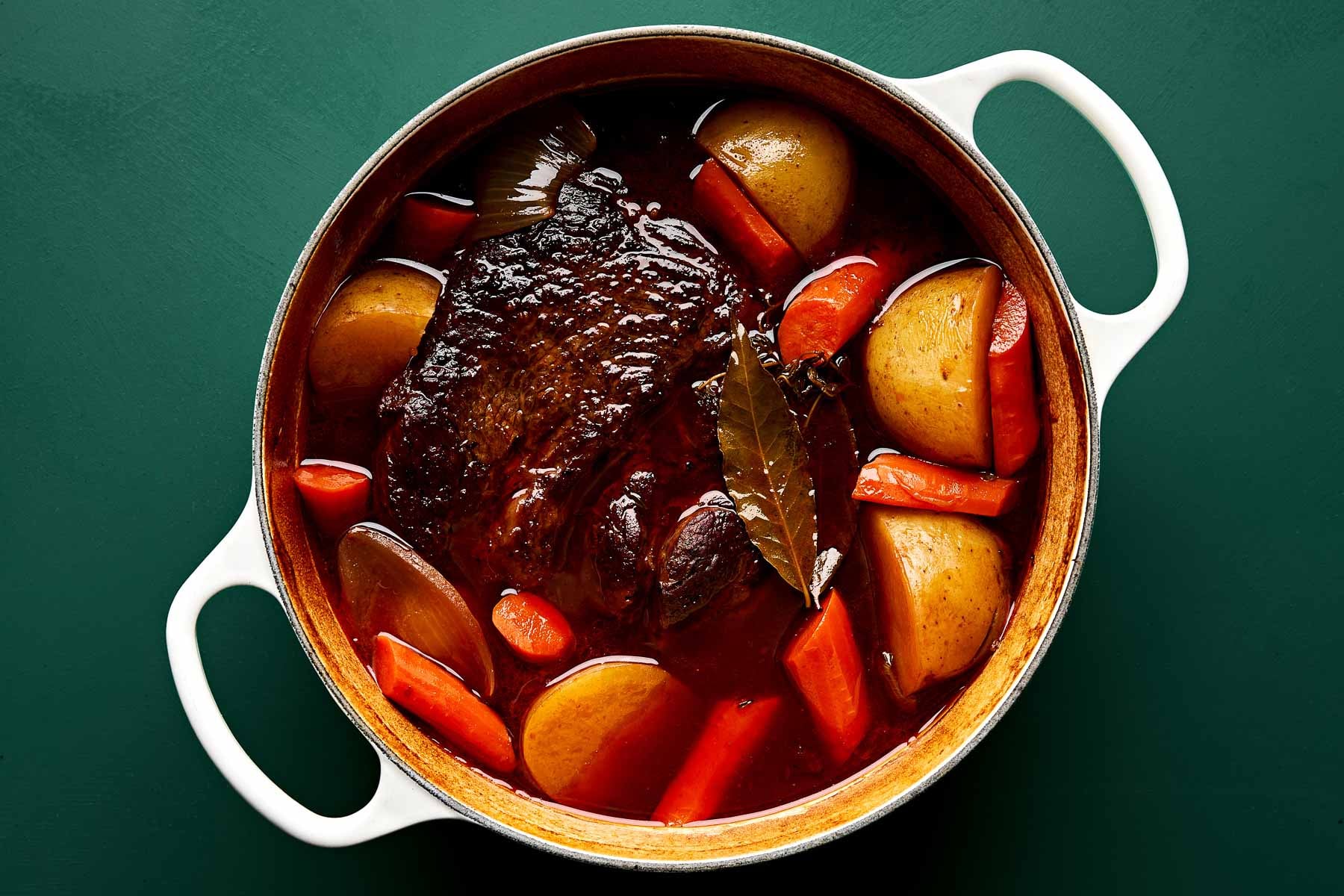 A white pot filled with beef pot roast, carrots, potatoes, onions, and bay leaves in a rich brown broth, viewed from above on a dark green background.