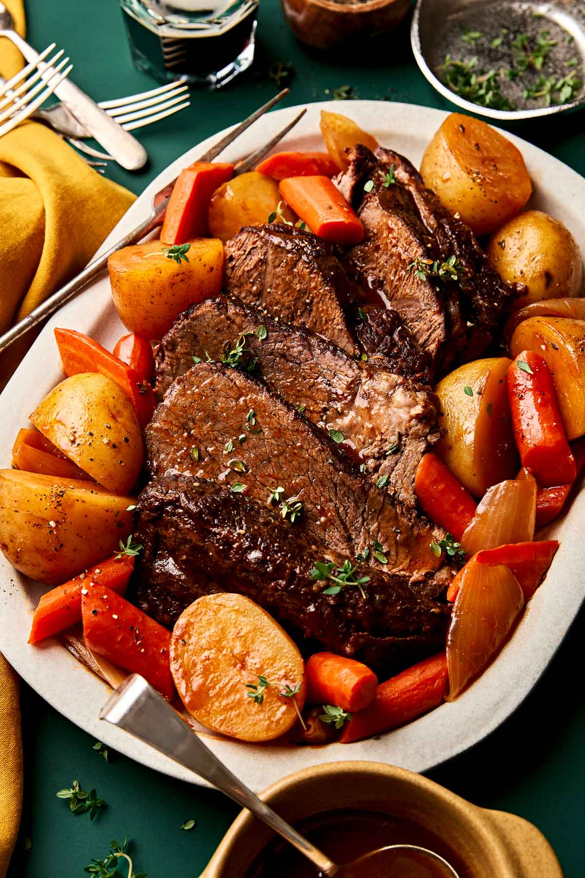 Guinness Pot Roast with Gravy A platter of pot roast with slices of tender beef, roasted potatoes, carrots, and onions, garnished with fresh herbs, served with a fork and surrounded by utensils and a yellow napkin.