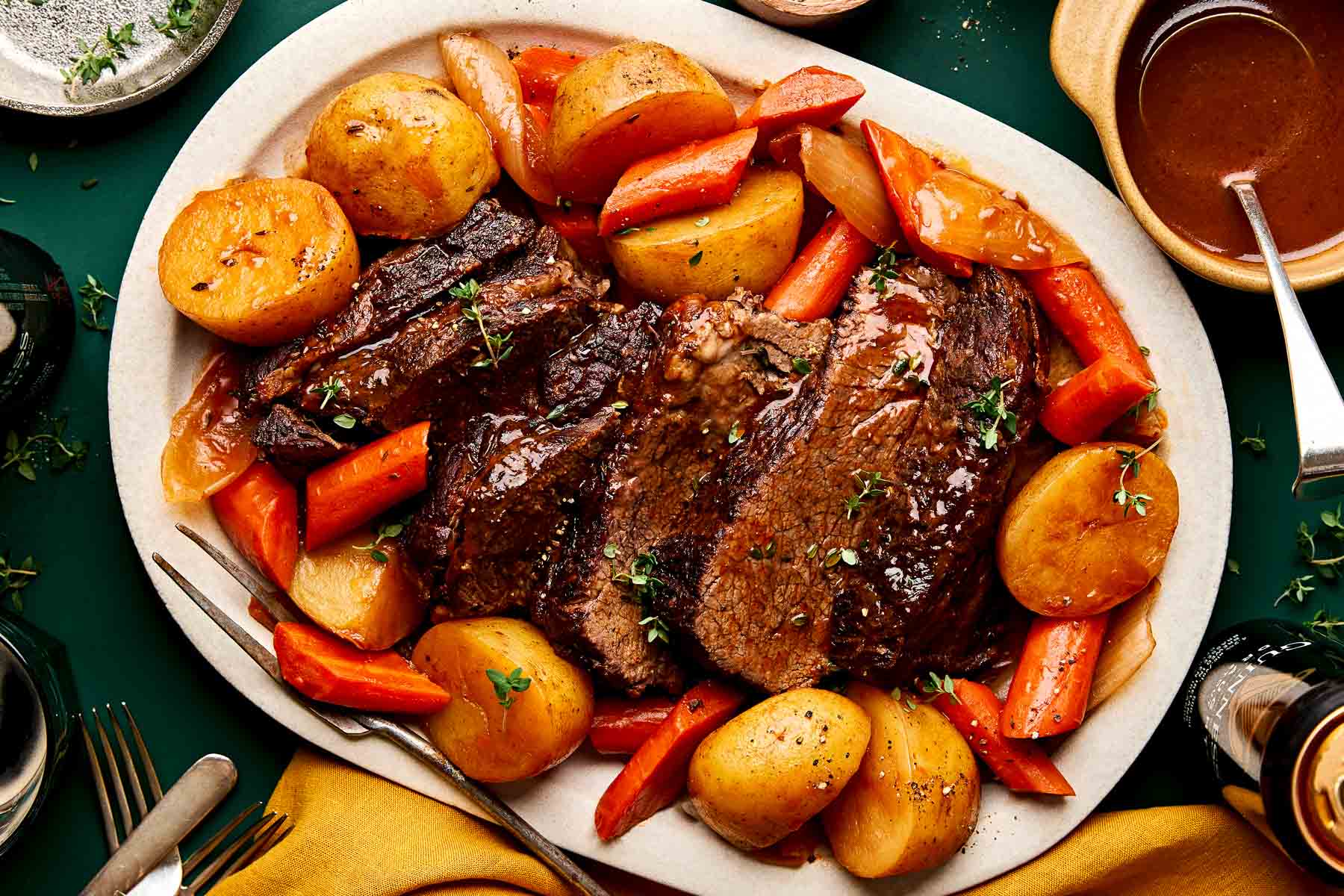 A white platter filled with sliced pot roast, whole potatoes, and carrots, all garnished with fresh herbs and served with gravy on the side.